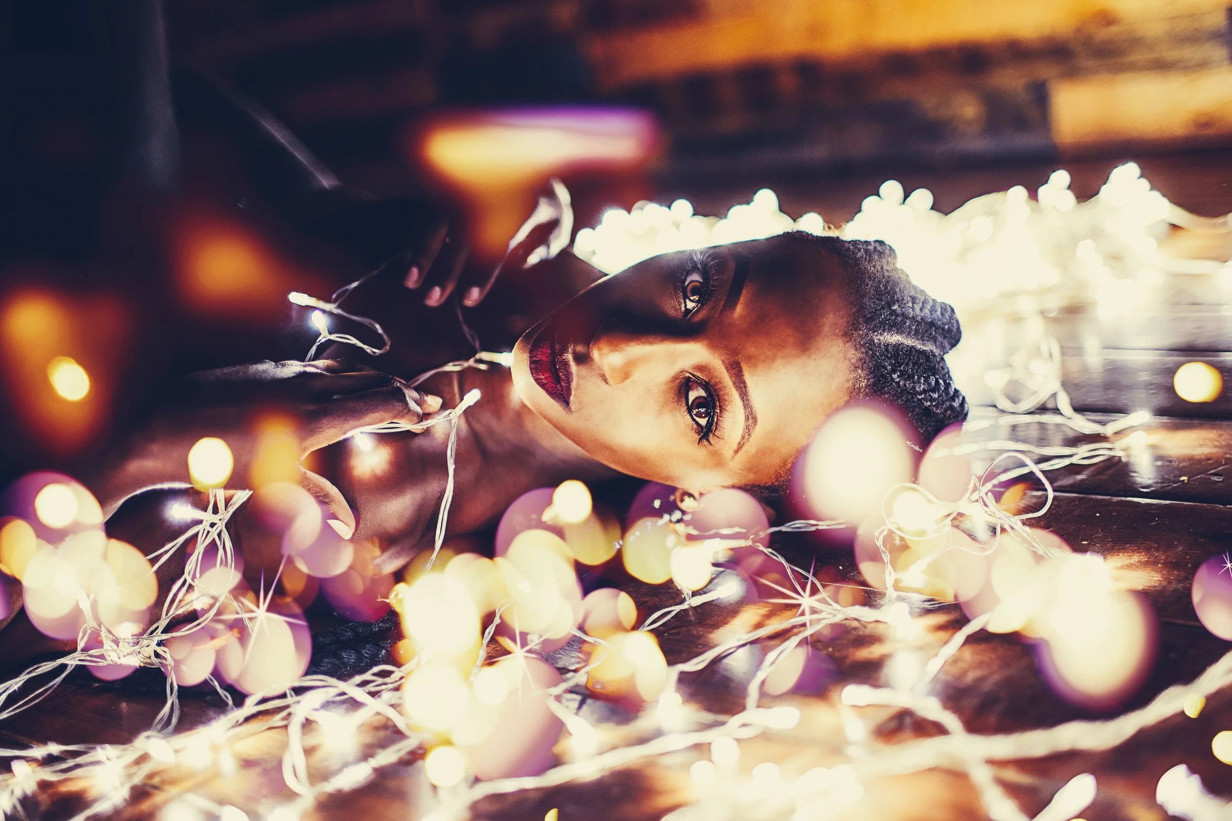 A woman lying on a wooden floor surrounded by colorful string lights, with a soft, warm glow illuminating her face and creating bokeh effects.
