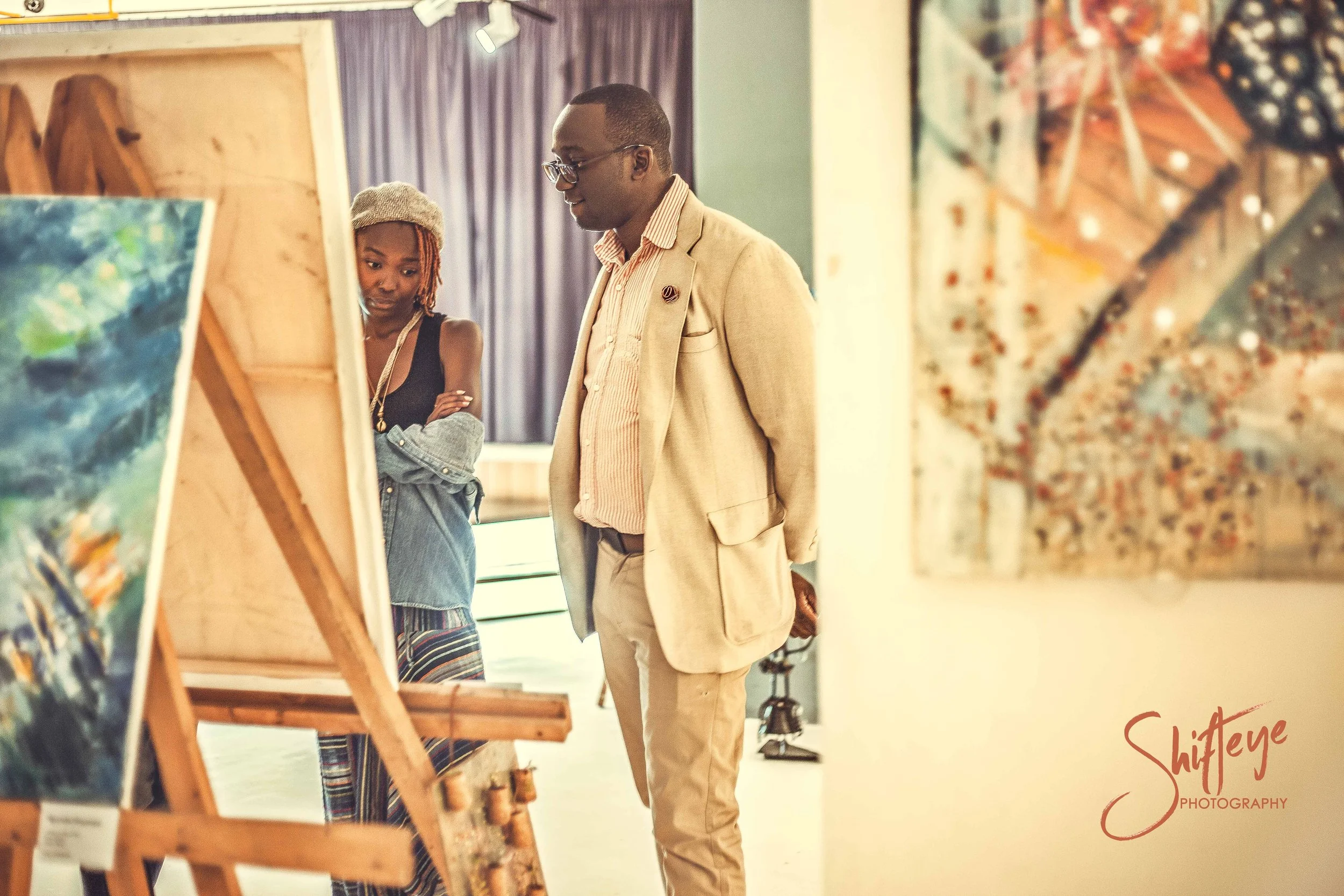 A man and a woman viewing artwork in an art gallery. The woman is wearing a gray beanie, black top, and striped pants, while the man is dressed in a beige blazer and pants. Both appear to be engaged in observing the paintings.
