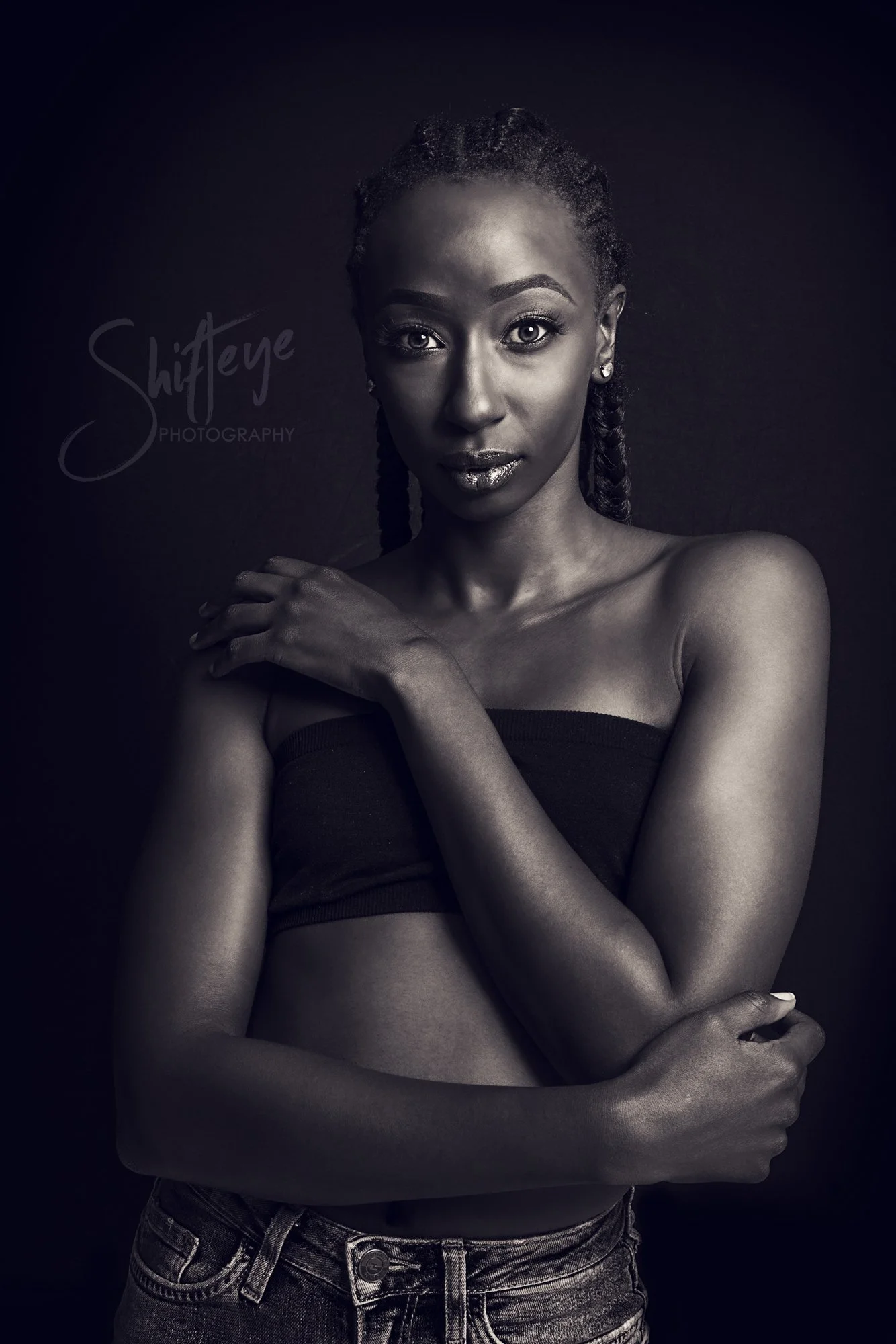 Black and white portrait of a woman with braided hair, wearing a strapless top and jeans, standing against a dark background, looking directly at the camera.