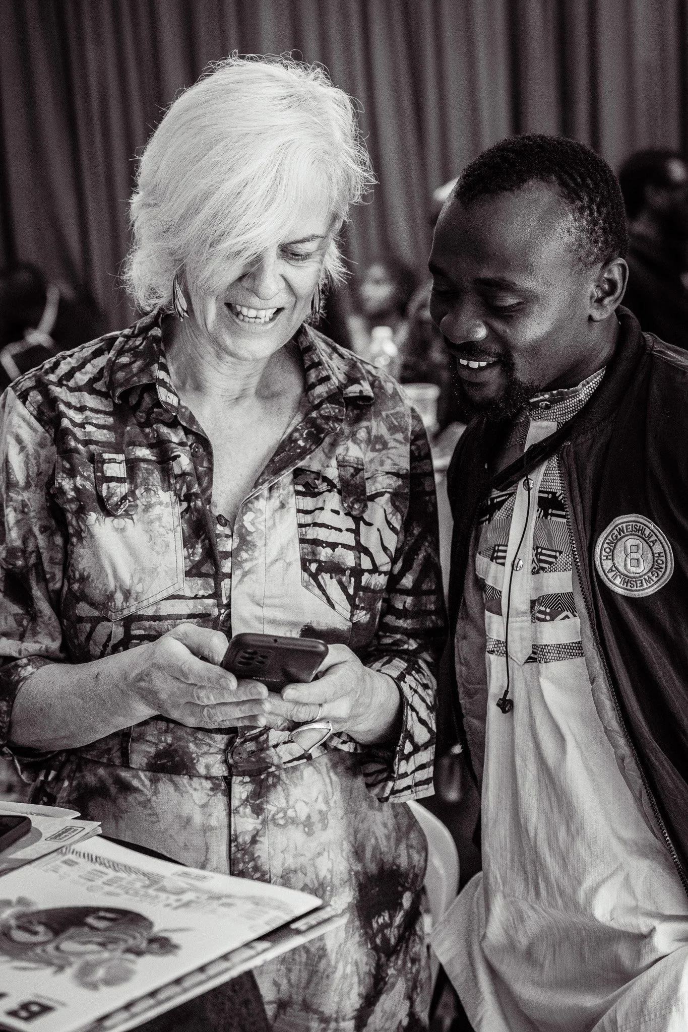 Two people, an older woman with white hair and a young man with short dark hair, are smiling and looking at a smartphone together at a gathering. There are other people blurred in the background.