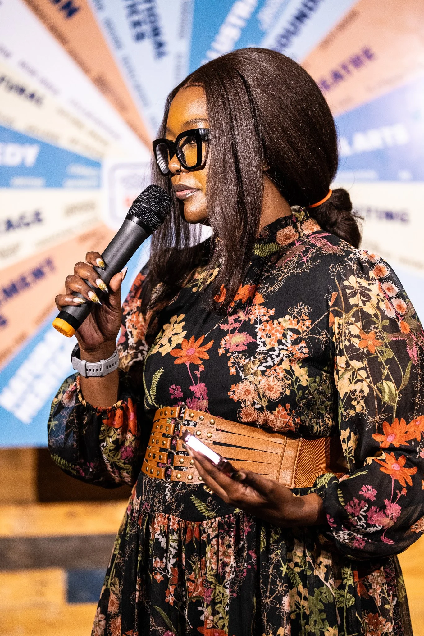 A woman with dark skin and long, dark hair wearing glasses, a floral dress, and a tan belt, holding a microphone and a smartphone, speaking at an event with a colorful background.