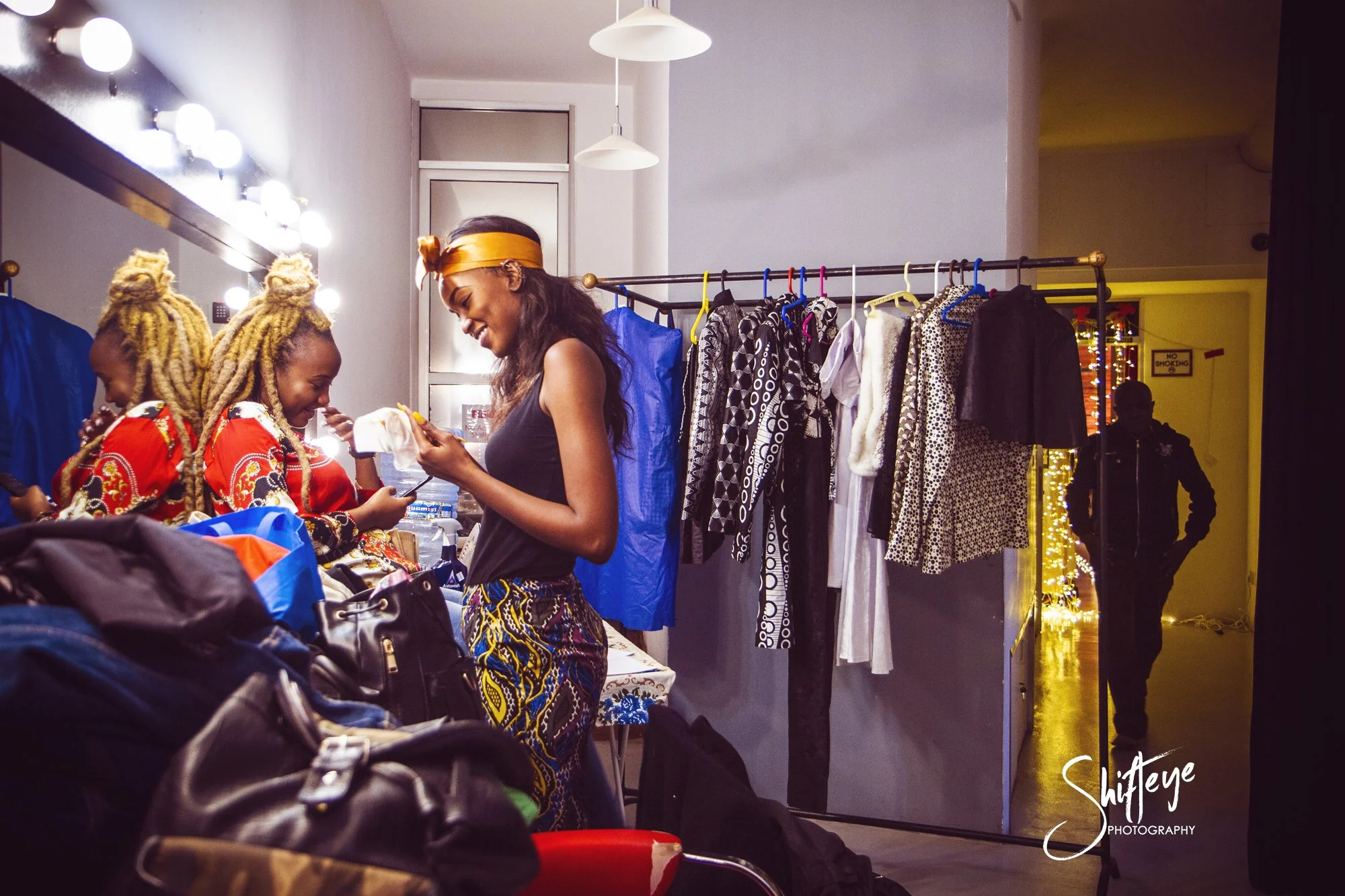 Two women with blonde dreadlocks, wearing colorful clothing, are smiling and talking to a woman with long dark hair, dressed in black, inside a dressing room with bright mirror lights. There are clothes on a rack behind them and bags on a table in fr