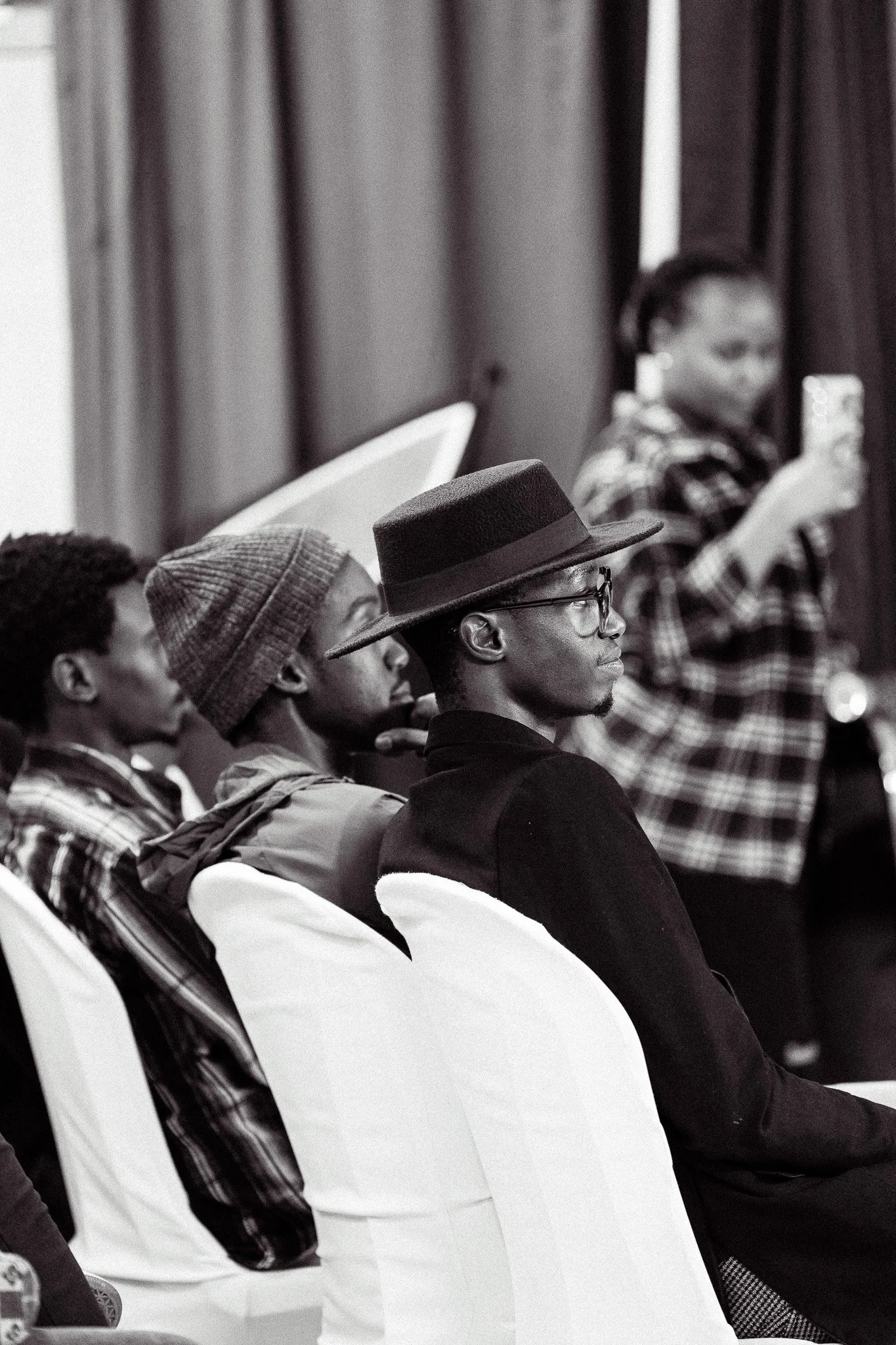 Black and white photo of three young men sitting in chairs and a young woman standing in the background, using a smartphone. The young men are focused on a presentation or event, with one wearing glasses and a wide-brim hat, another with a beanie, an