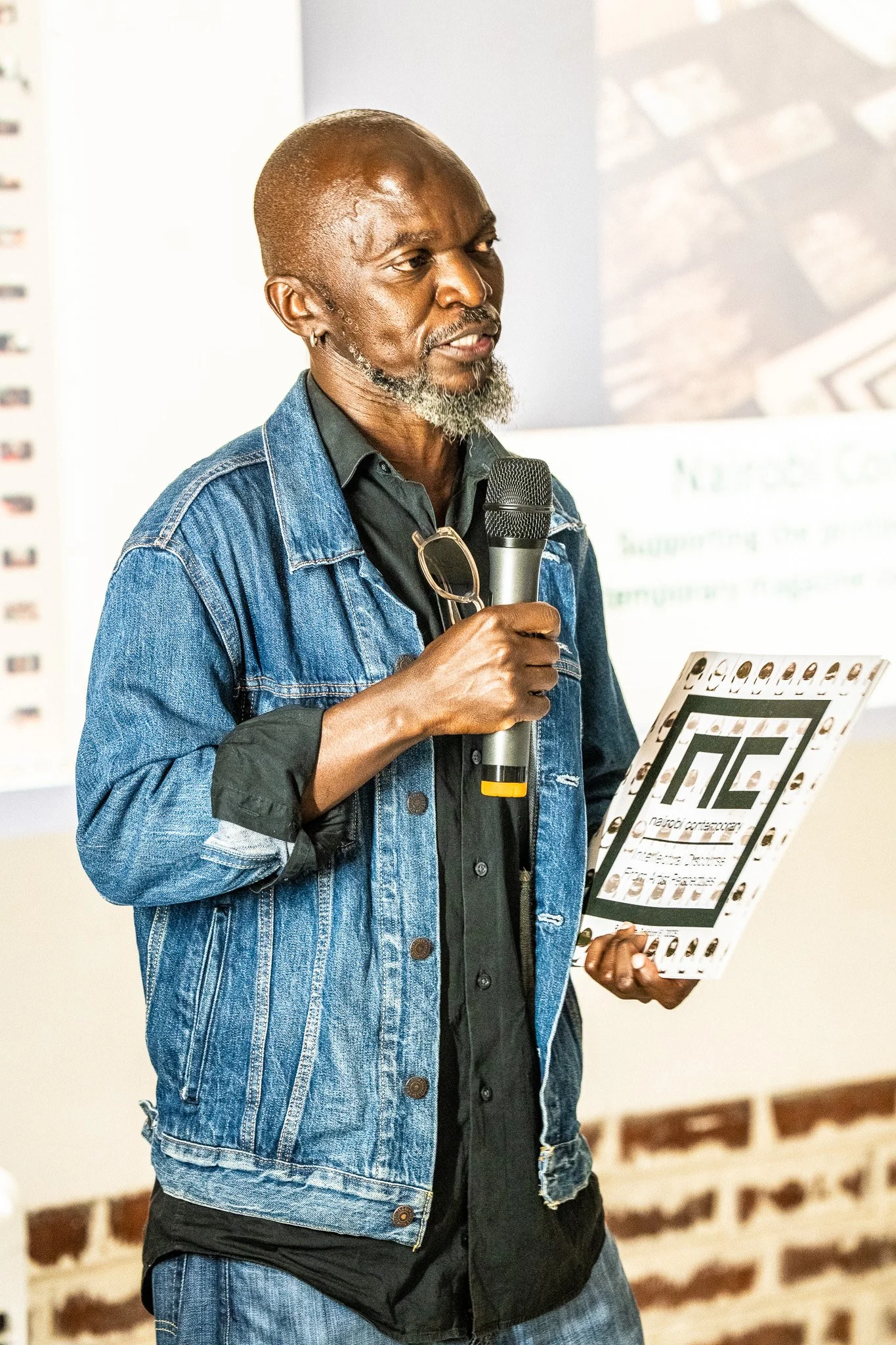 A man with a gray beard holding a microphone and a presentation board during a presentation.