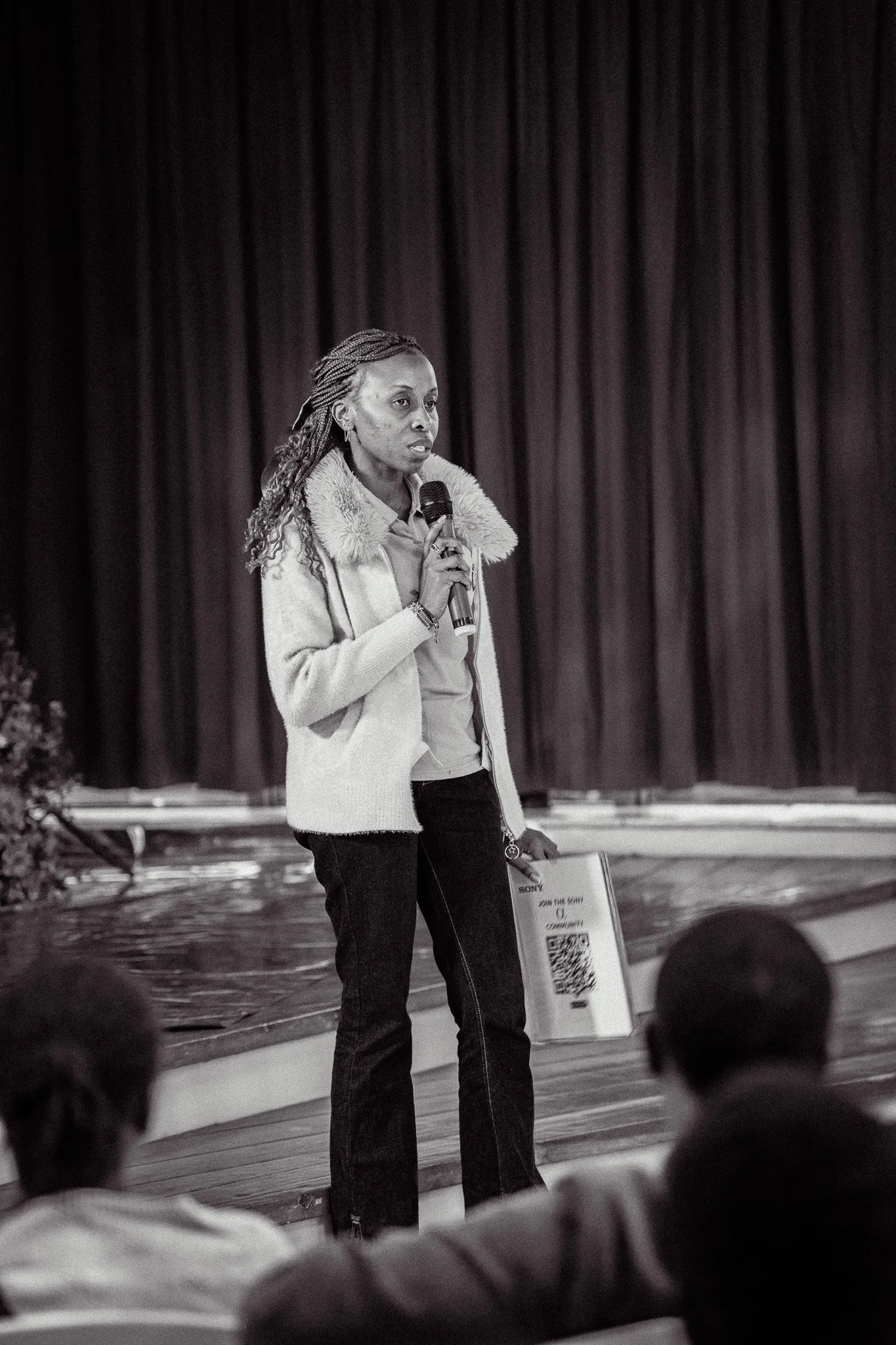 A woman speaking into a microphone on stage during a presentation or speech, with an audience in the foreground, black and white photo.