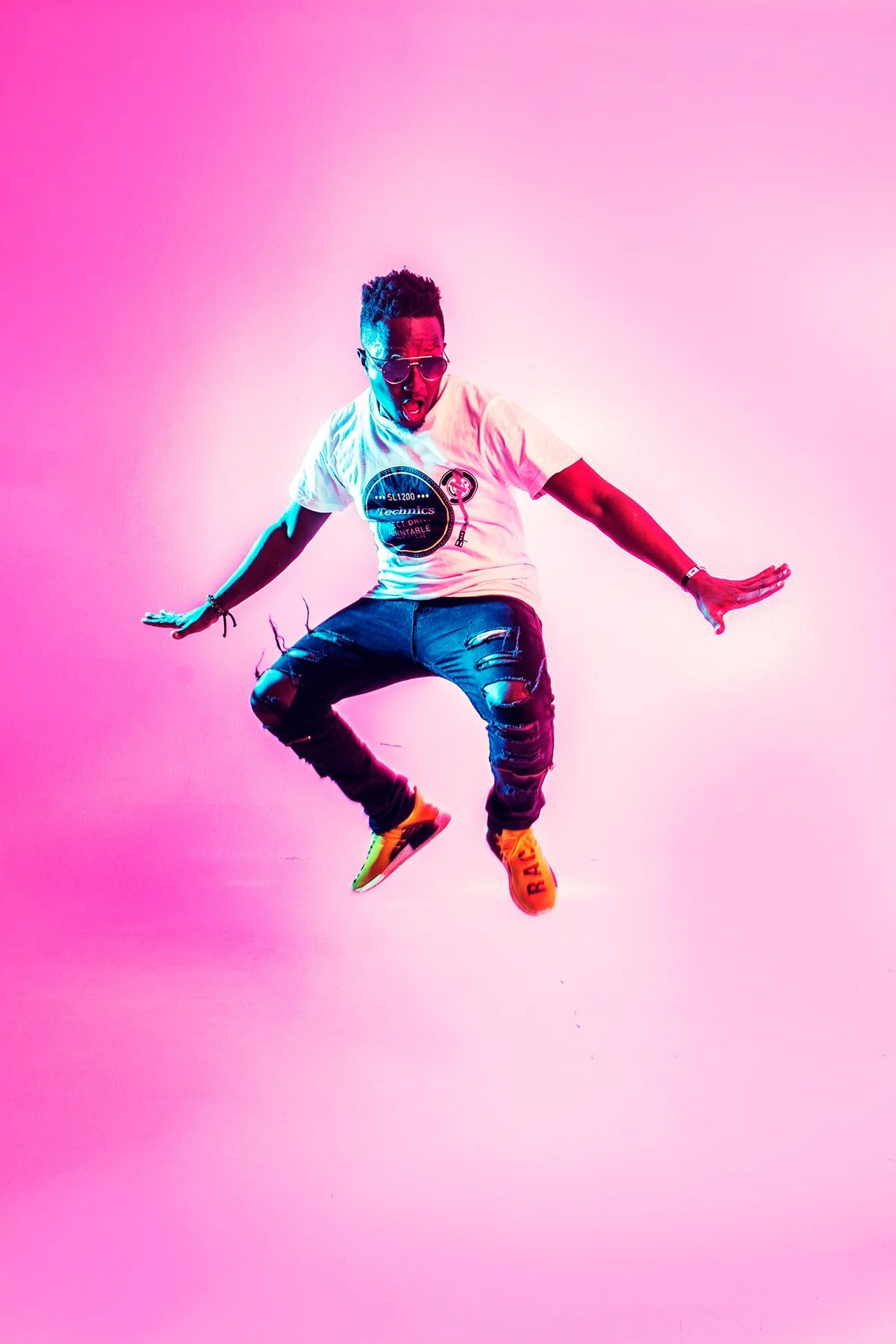 A young man wearing sunglasses, a white T-shirt, ripped jeans, and colorful sneakers is jumping against a pink background with a vibrant, neon glow.