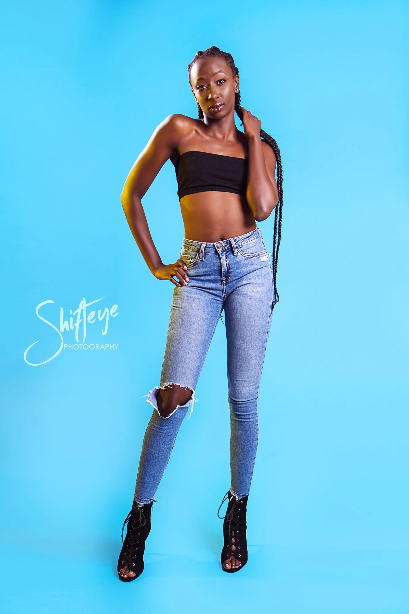 A woman standing against a bright blue background, wearing a black bandeau top, ripped jeans, and black lace-up high heels, with her left hand on her hip and her right hand touching her neck.