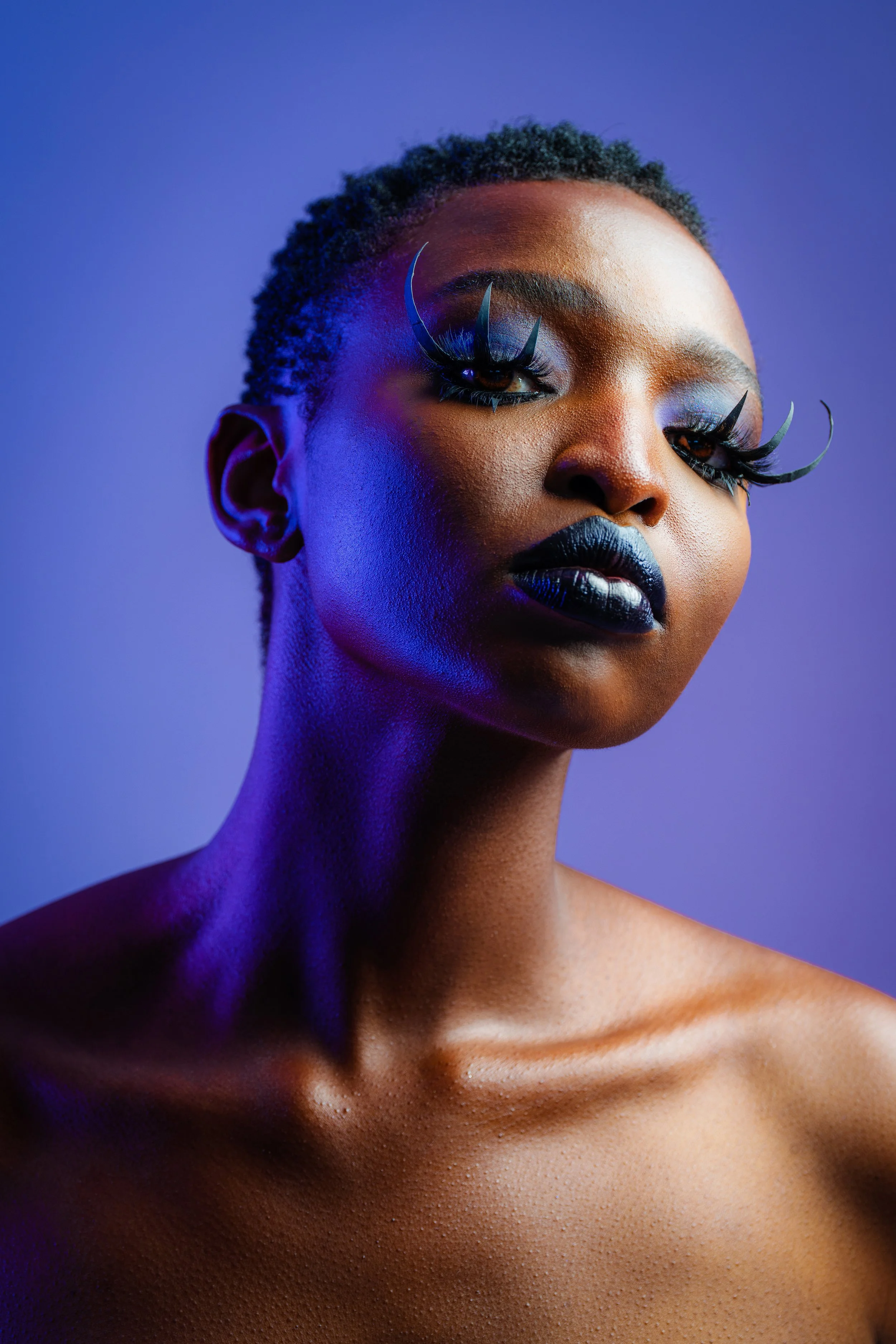 Portrait of a woman with bold makeup, eye lashes, and metallic lips, against a purple background.