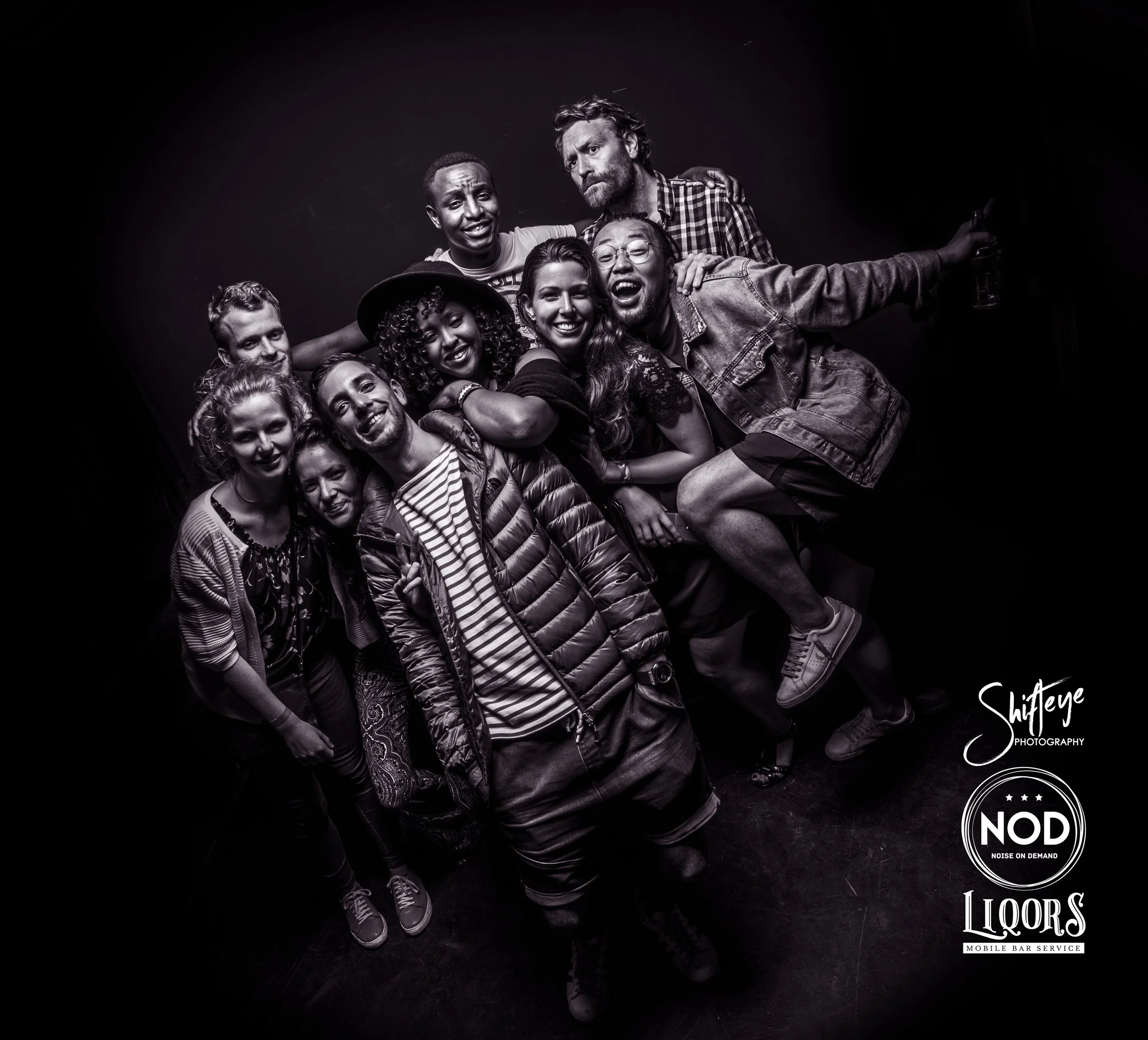 Group of people smiling and posing together in a black-and-white photo, some holding drinks, with logos for Shifteye Photography, NOD Noise on Demand, and LOORS Mobile Bar Service in the bottom right corner.