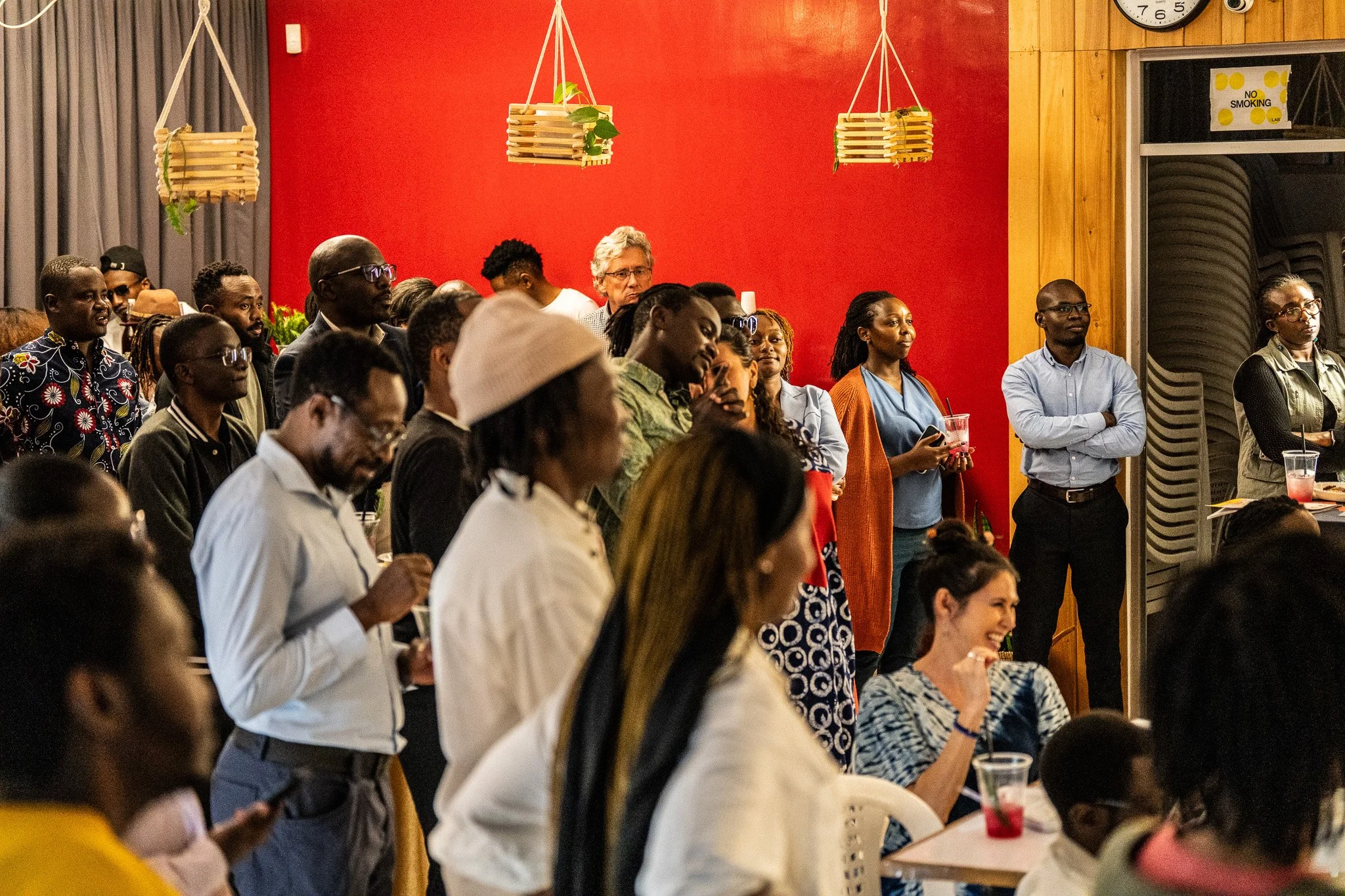 People gathered at an indoor event, standing and seated, with a red wall and wooden paneling in the background.