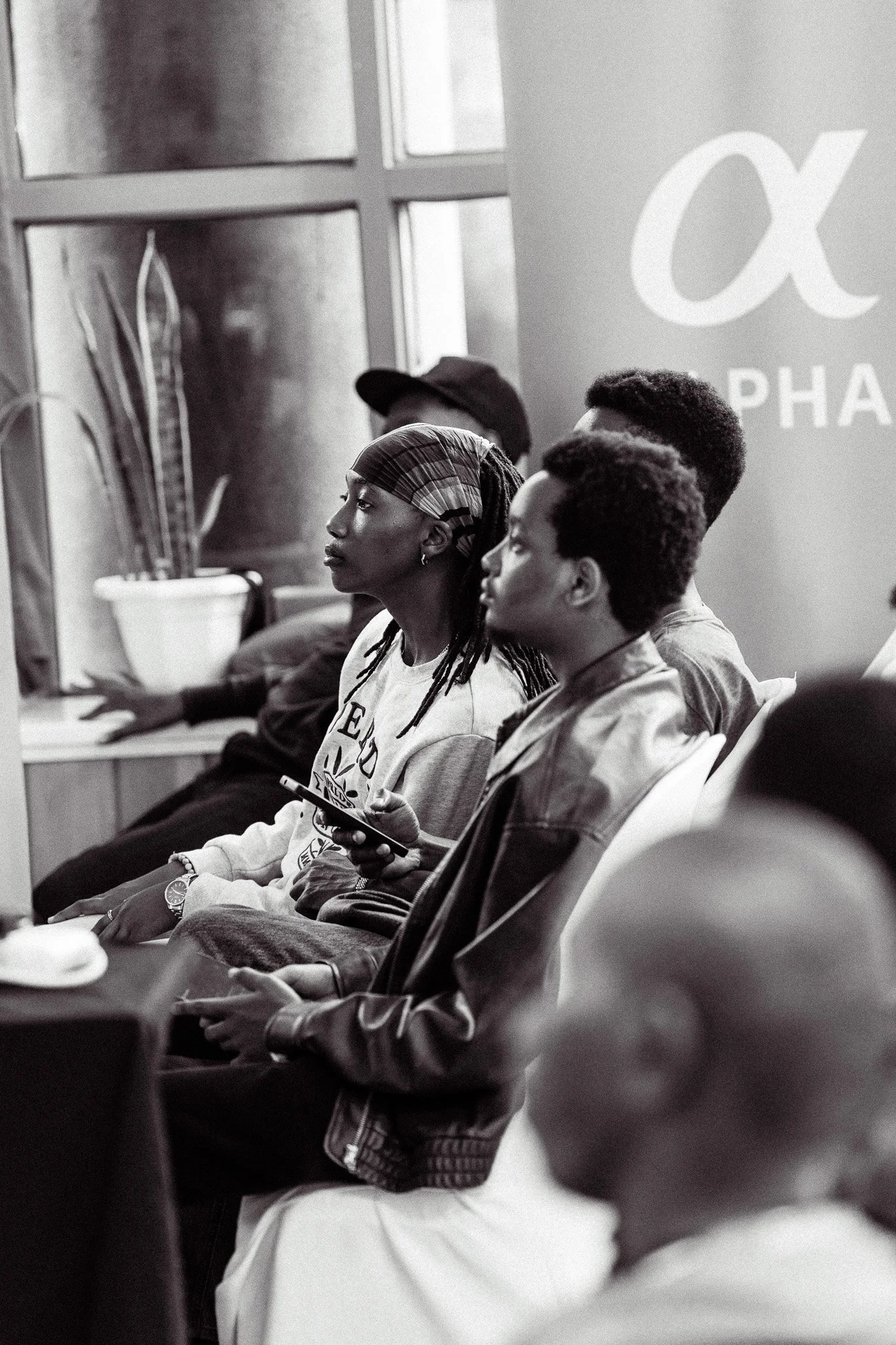 A group of diverse individuals sitting in a conference room, attentively listening with some using their smartphones, with a large alpha symbol and the word 'Alpha' on a banner in the background.