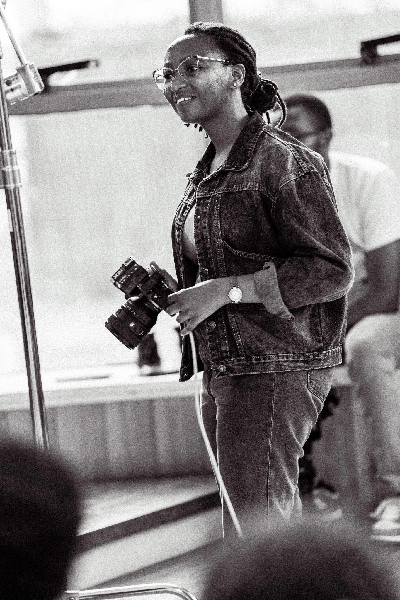 A woman with glasses and dreadlocks, wearing a denim jacket and jeans, holding a camera on a film set or studio.