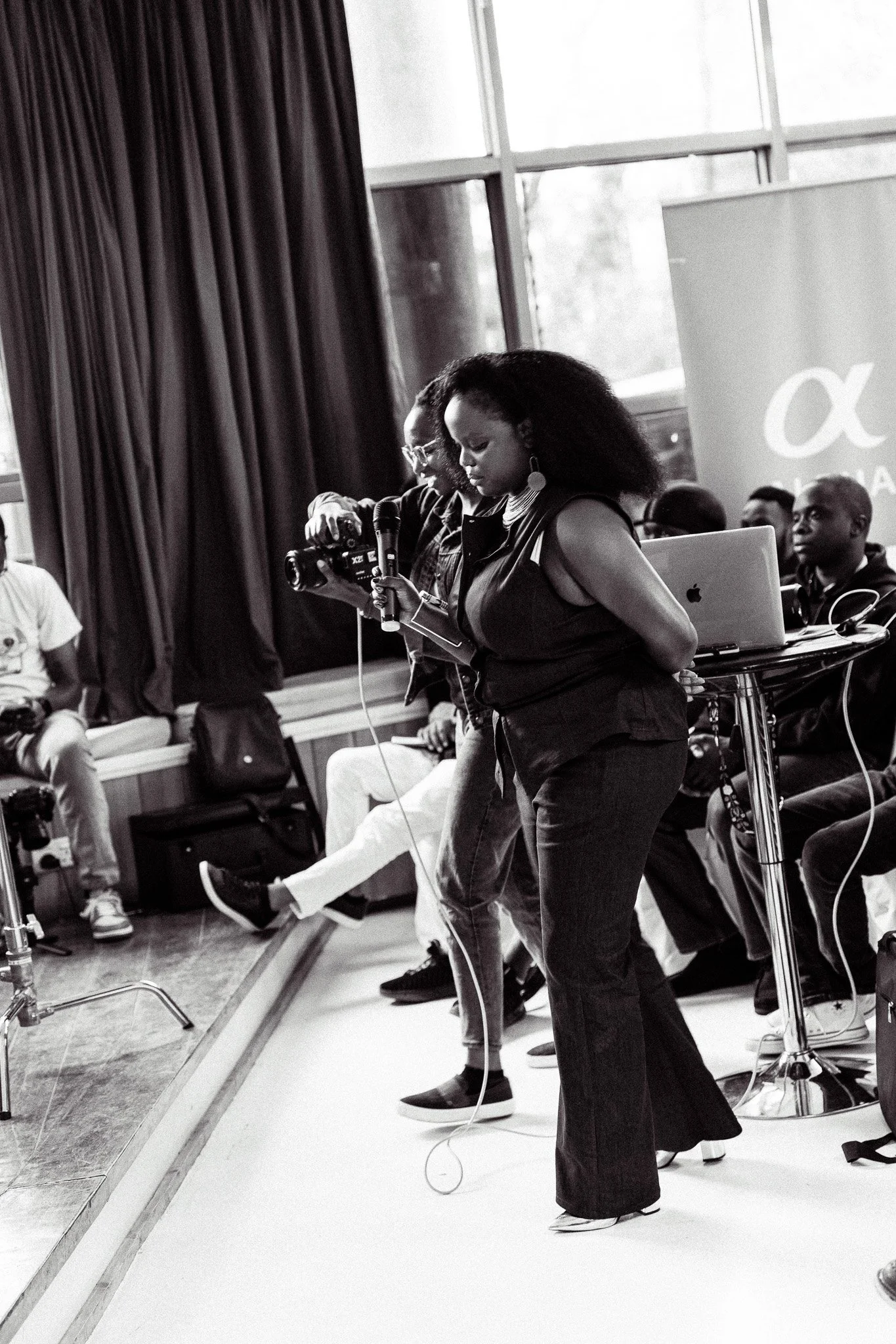 A woman with large curly hair and earrings, holding a phone, stands at a presentation or event with a group of people seated behind a table with a laptop. The scene is indoors with large windows and curtains.