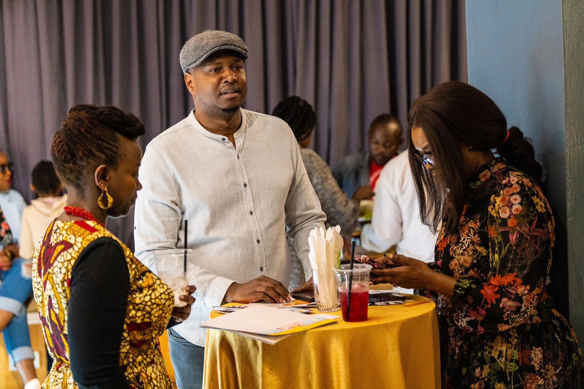 People standing at a table during a social event, with drinks and papers, engaged in conversation.