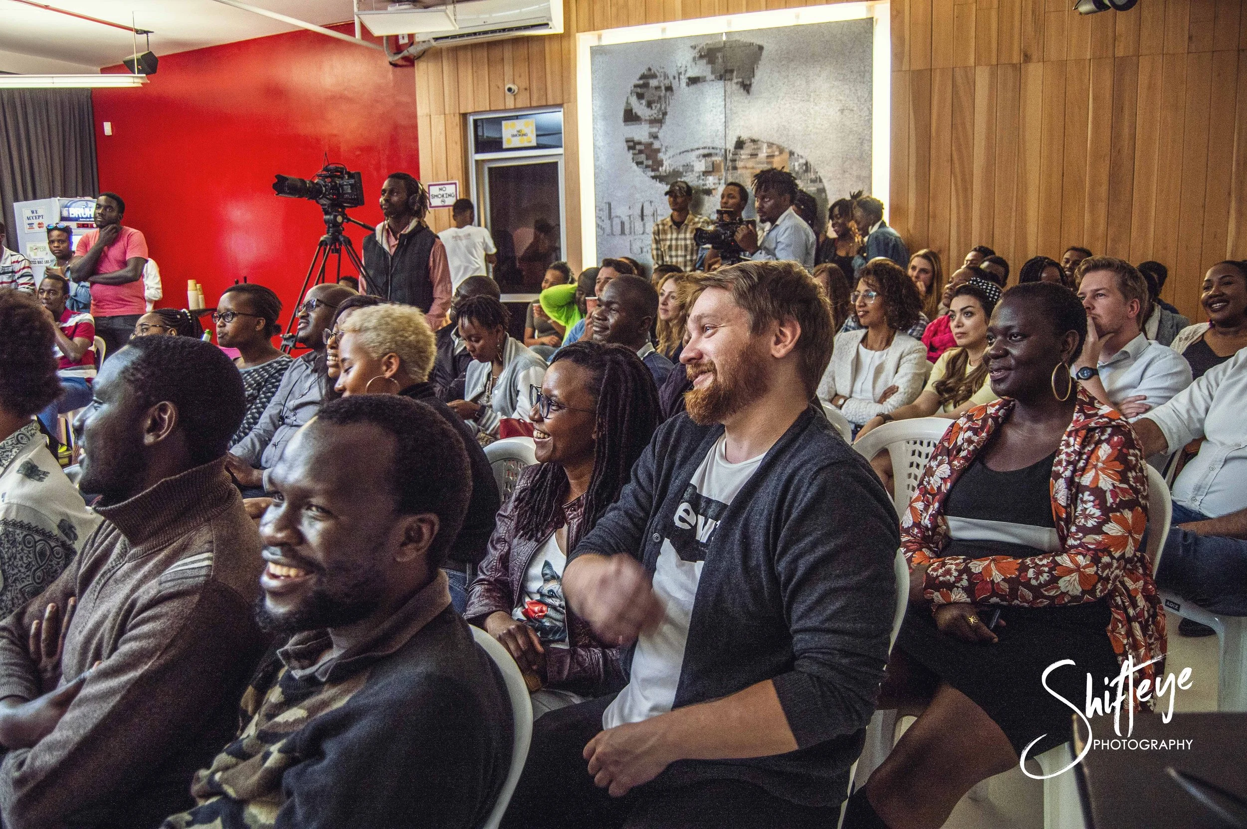 People seated in an audience at an indoor event, smiling and enjoying a presentation or performance. Some are looking towards a stage or speaker, with a cameraman recording in the background. The room has red and wooden walls, and a diverse group of 