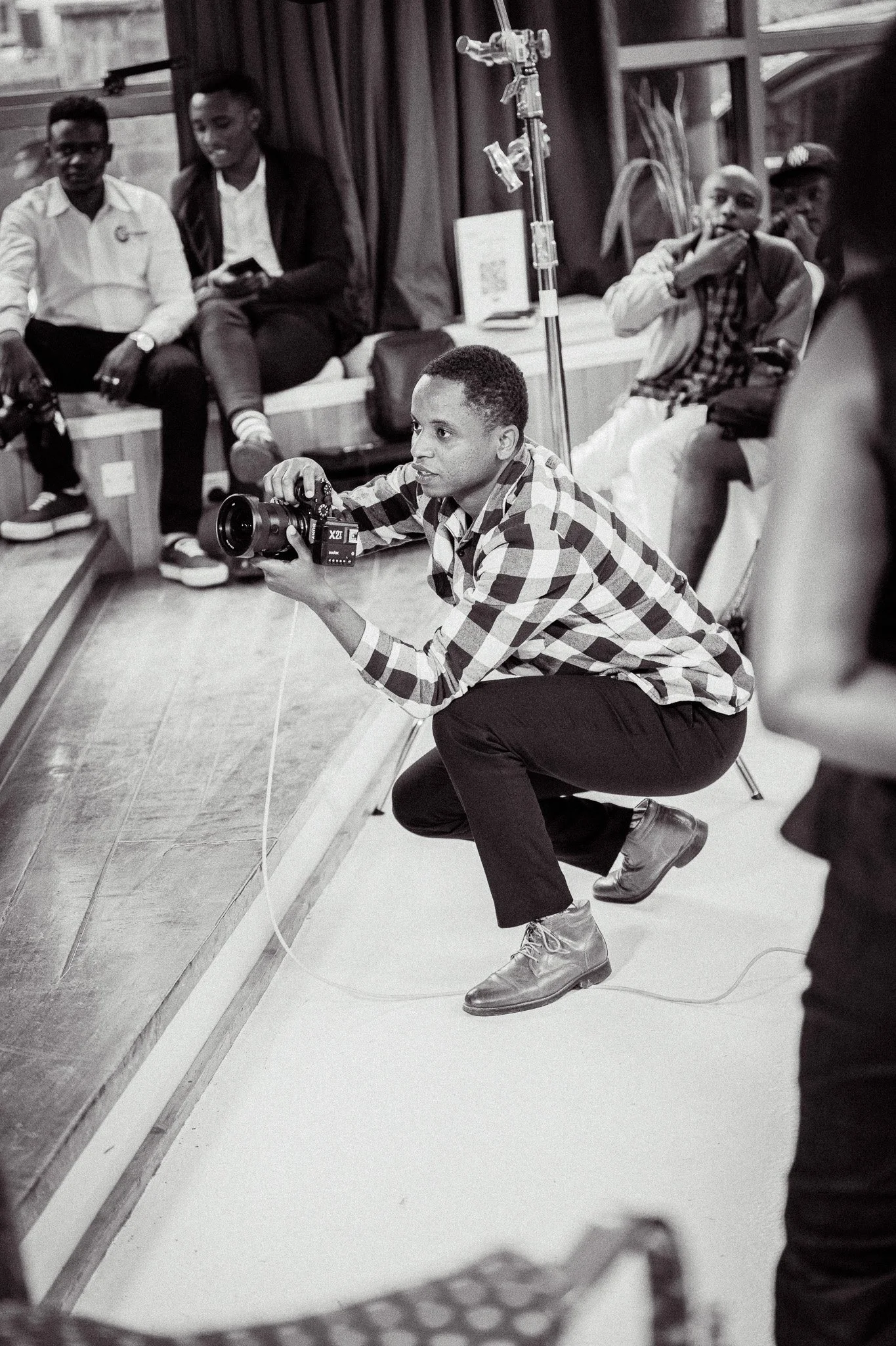 A photographer squatting on the floor, holding a camera, taking photos at an indoor event with seated attendees in the background.