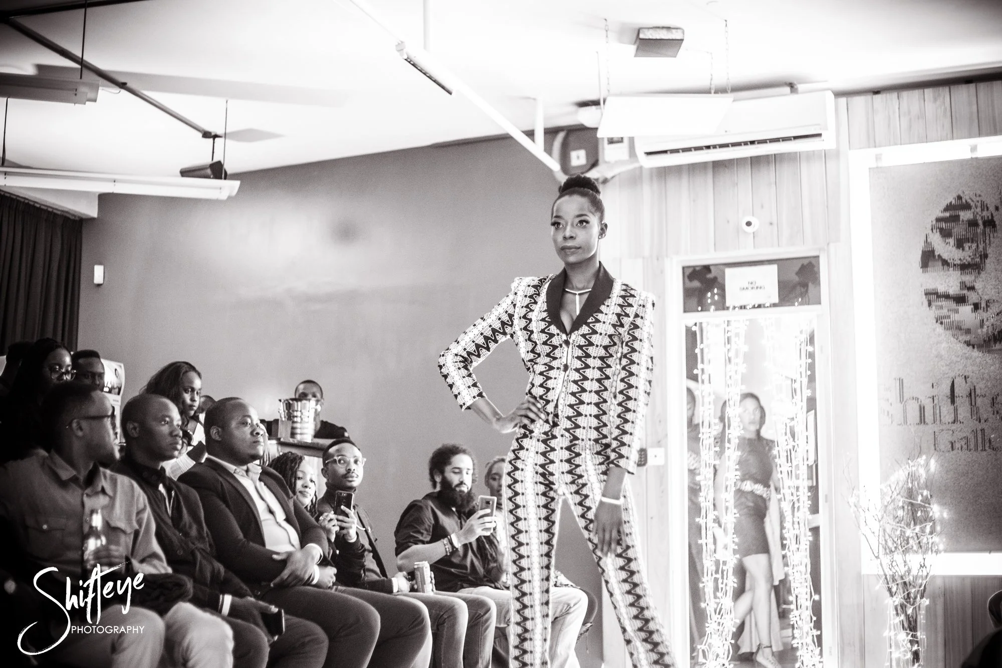 A woman walking on a runway during a fashion show, wearing a patterned suit. The audience is seated on the side, watching her.
