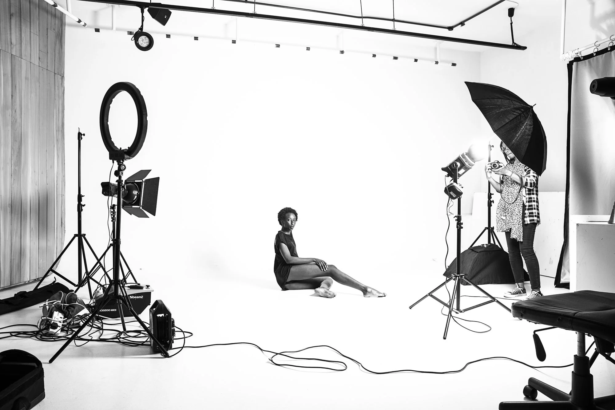 A woman sits on the floor of a photography studio, surrounded by professional lighting and equipment. A photographer with an umbrella and camera is taking her picture against a plain white backdrop.
