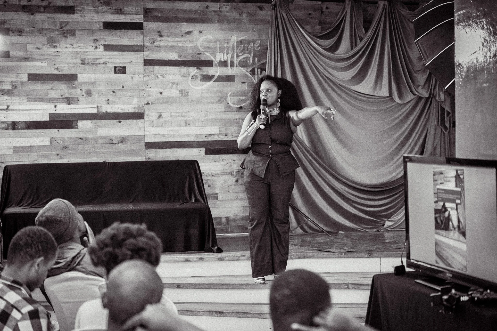 A woman speaking on stage with a microphone, gesturing with her right hand, in front of an audience, with a wood-paneled wall, a curtain, and a decorated background behind her.