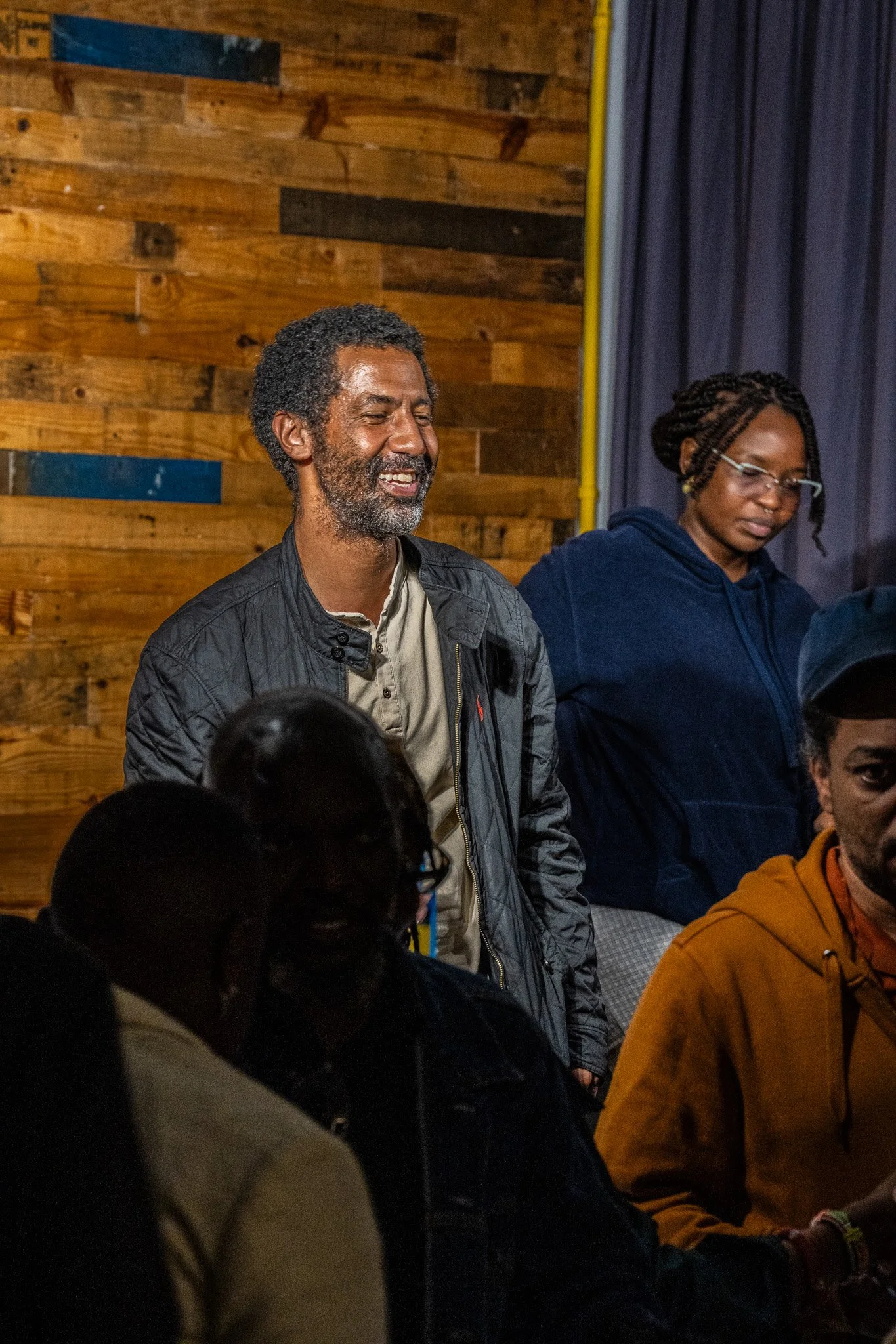 A group of people are gathered indoors, with two standing in focus. The man in the center has gray curly hair and a beard, wearing a tan shirt and a black jacket, smiling. The woman next to him has glasses, braids, and is wearing a dark hoodie, looki
