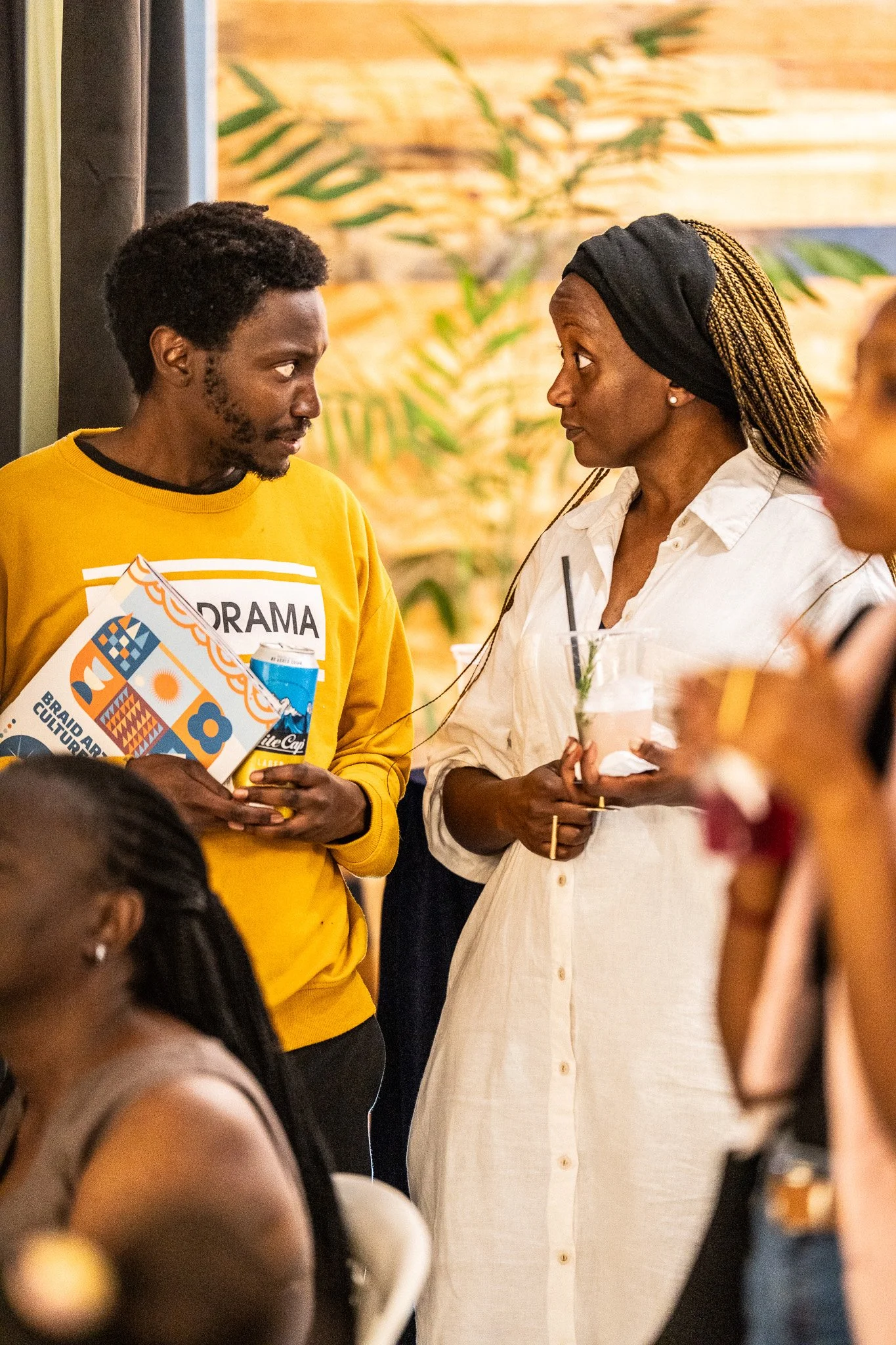 Two women are engaged in conversation at a social gathering, with one holding a drink and the other holding a booklet and a beverage in a plastic cup. The background features a wooden wall and greenery.