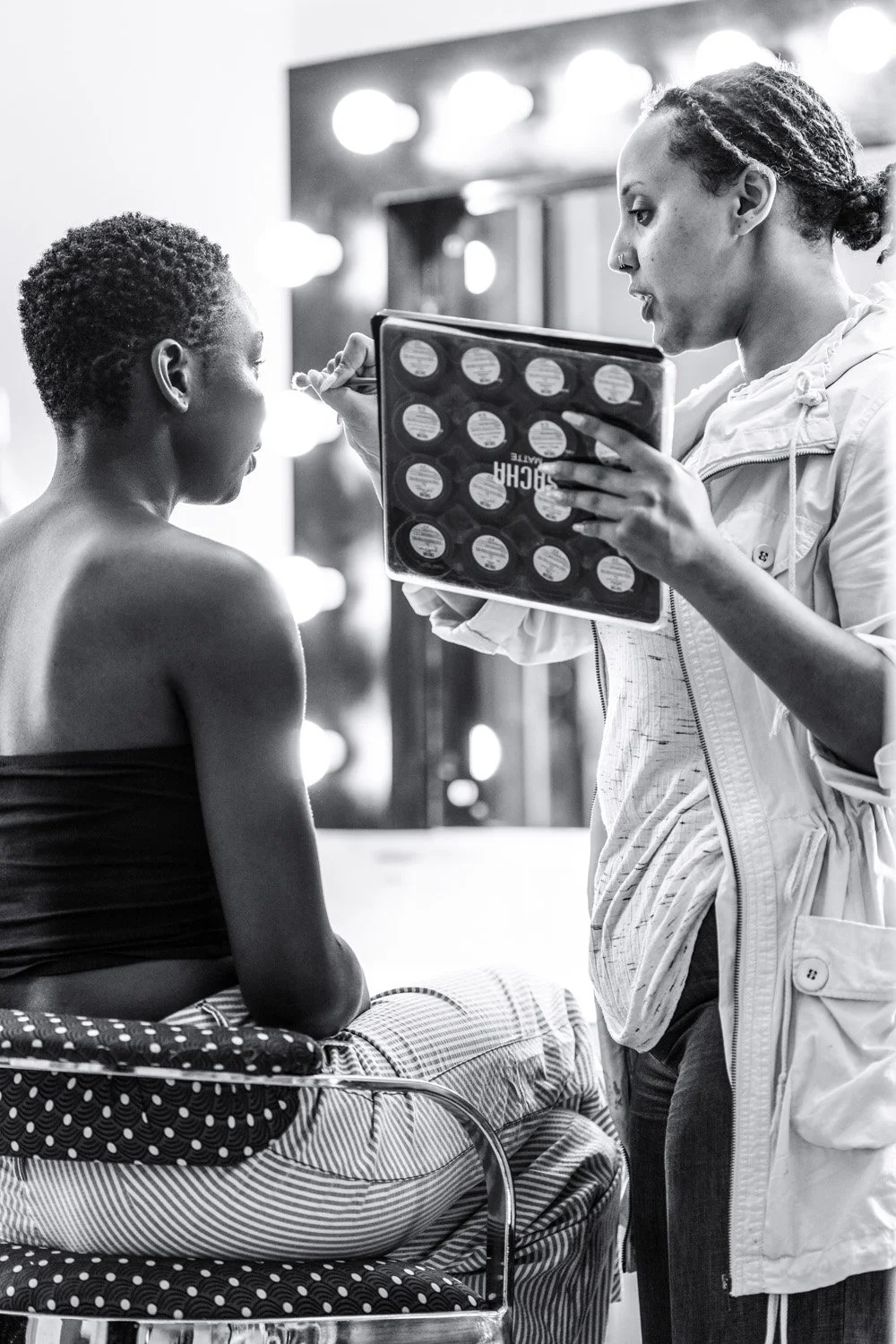 A woman is preparing to put makeup on a woman sitting in a wheelchair in a dressing room with illuminated vanity lights.