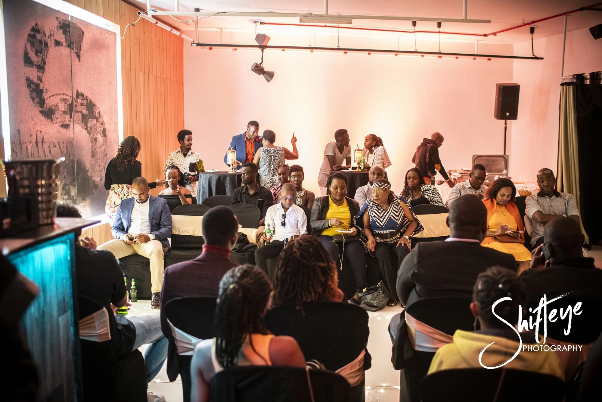 A group of people seated and standing at a social gathering or event in an indoor setting, with some at tables and others sitting in chairs, engaged in conversations and using their phones.