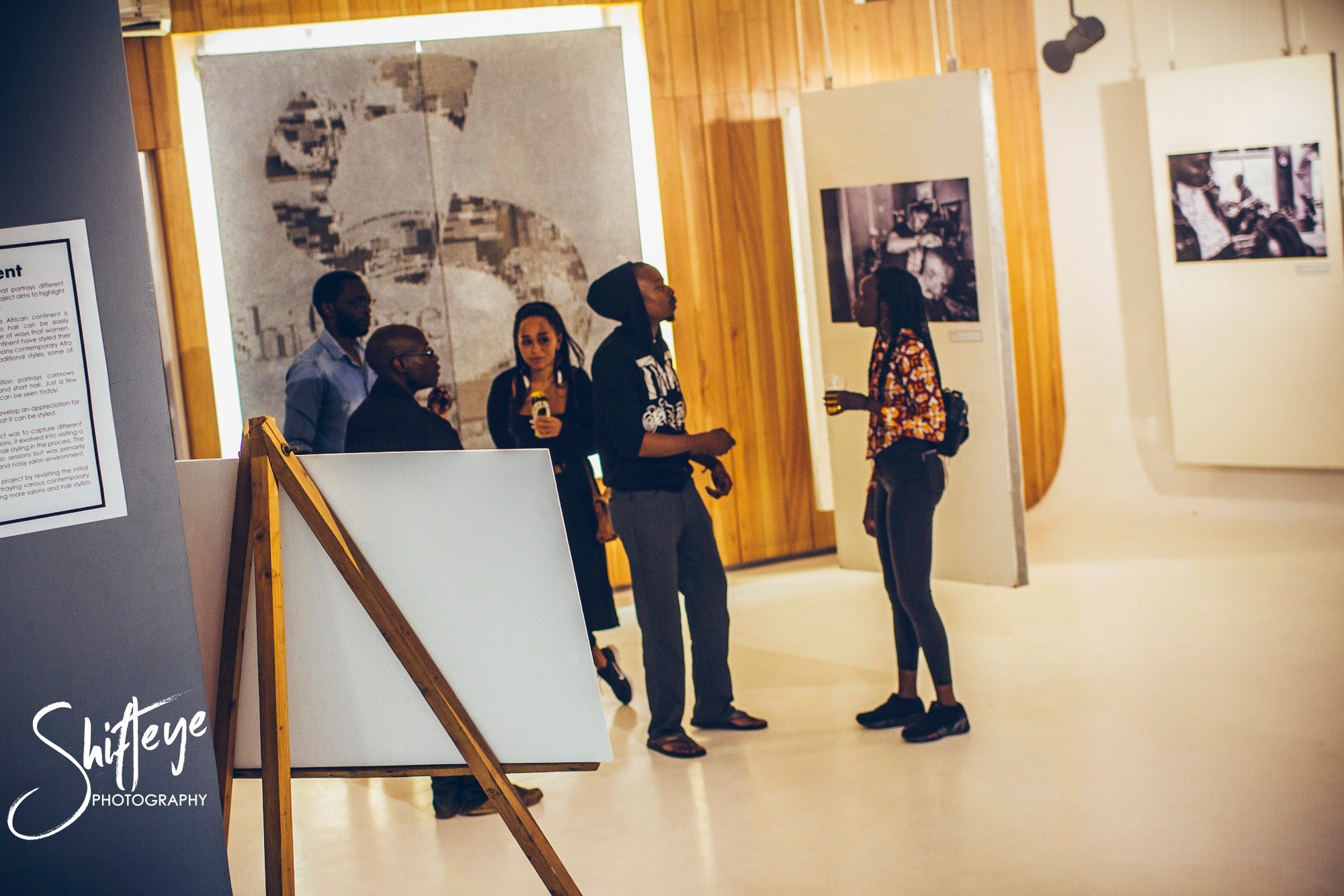 A group of five people inside an art gallery, observing and discussing paintings and photographs on display.