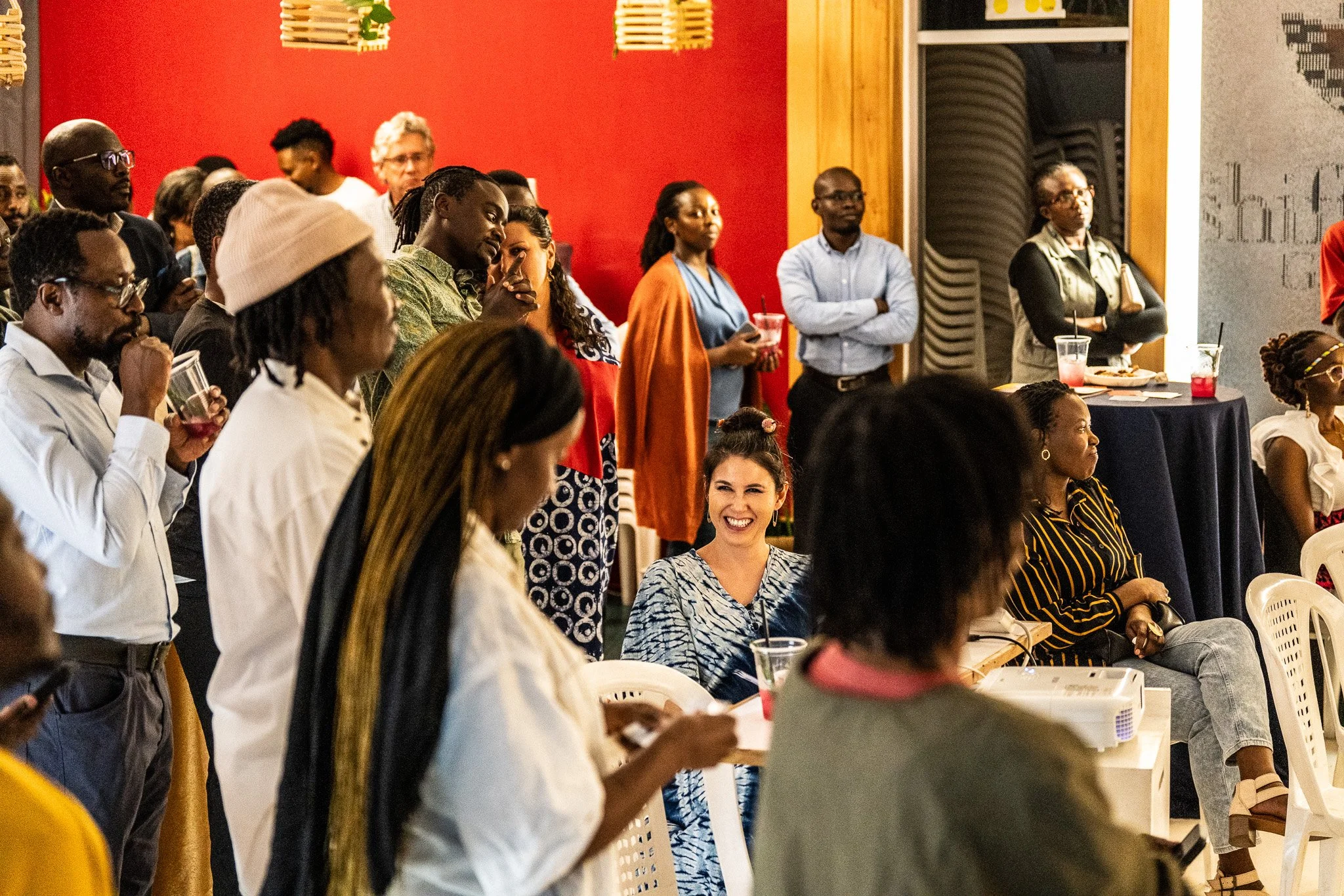 A group of diverse people at a social gathering, some standing and some seated, with drinks in hand, in an indoor setting with red walls and decorative lighting.