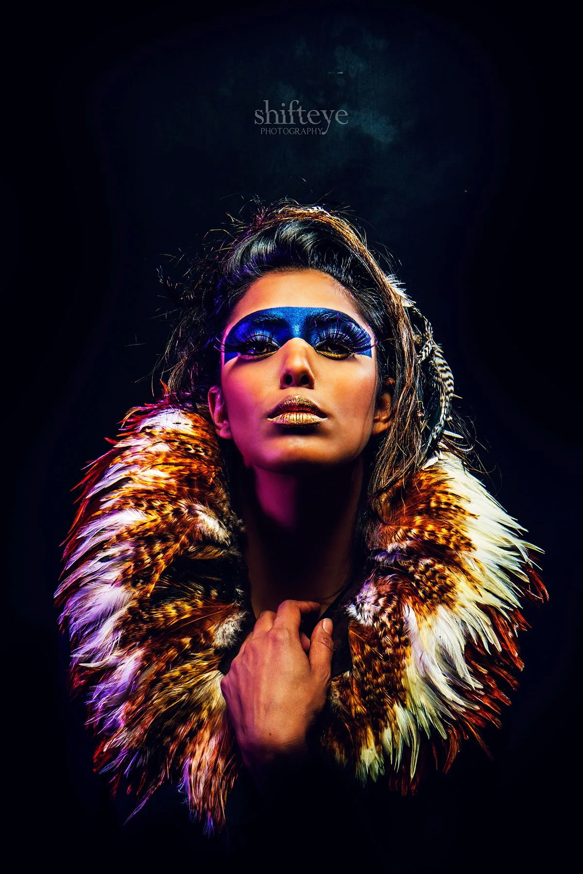 A woman with dramatic blue eye makeup and gold lipstick wearing a fur coat with leopard print pattern, posing with her hand near her chest against a dark background.