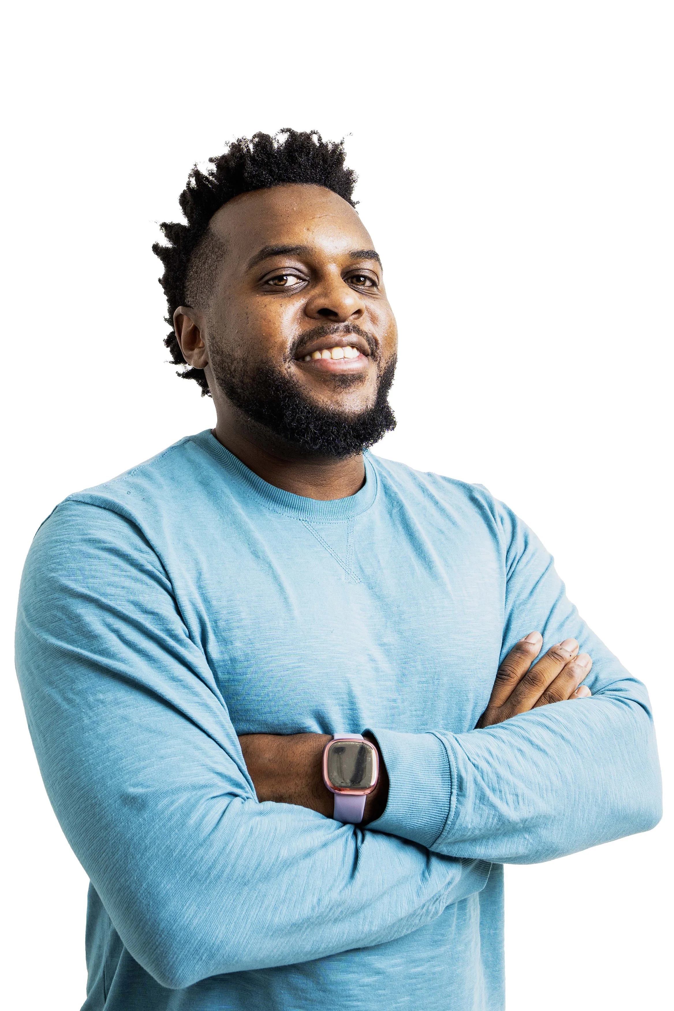 A man with dark curly hair and a beard wearing a teal long-sleeve shirt and a smartwatch, smiling with crossed arms.