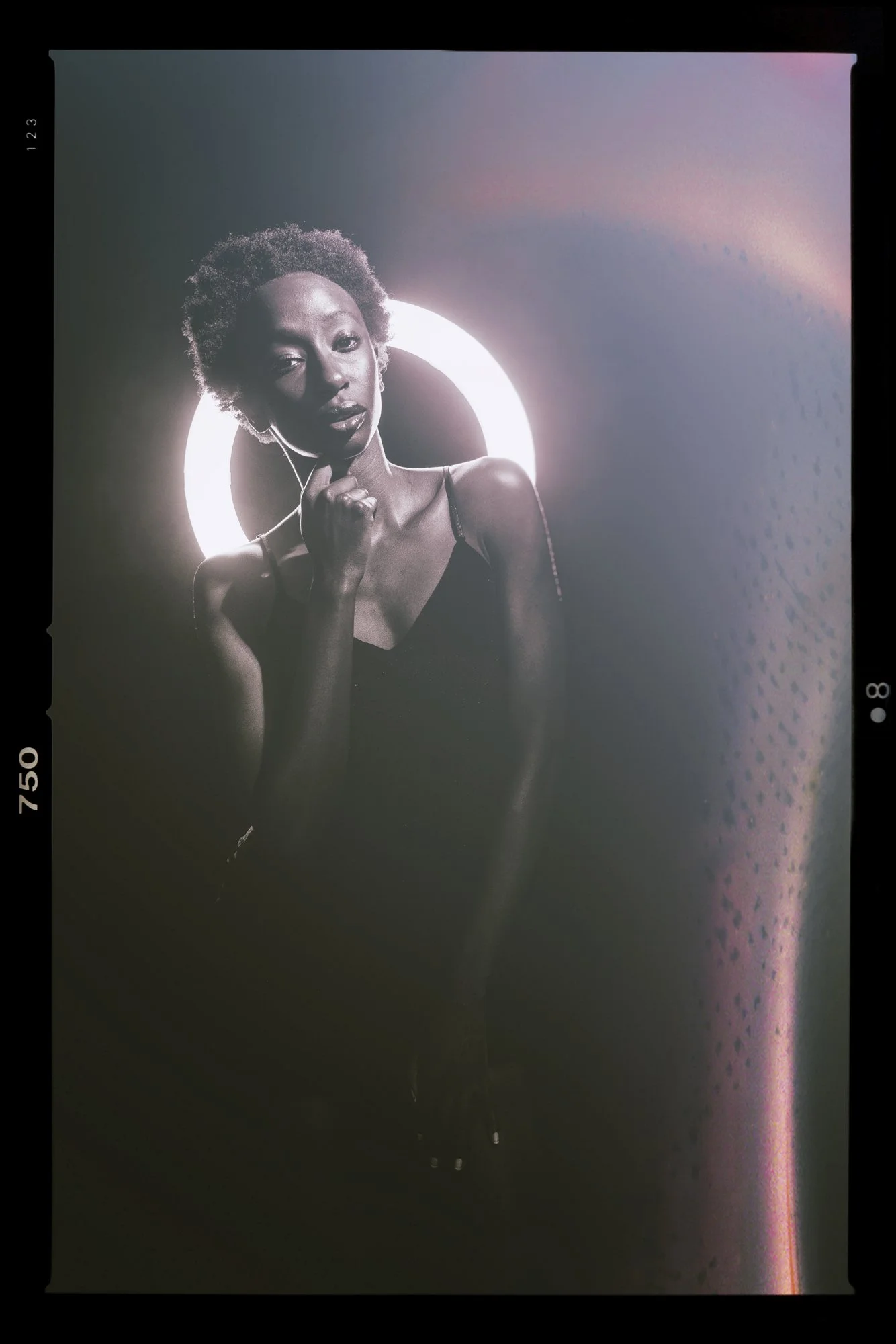 A black-and-white photograph of a woman with short curly hair, wearing a spaghetti strap top, standing in front of a glowing circular light. The woman has a contemplative expression, with one hand near her chin.