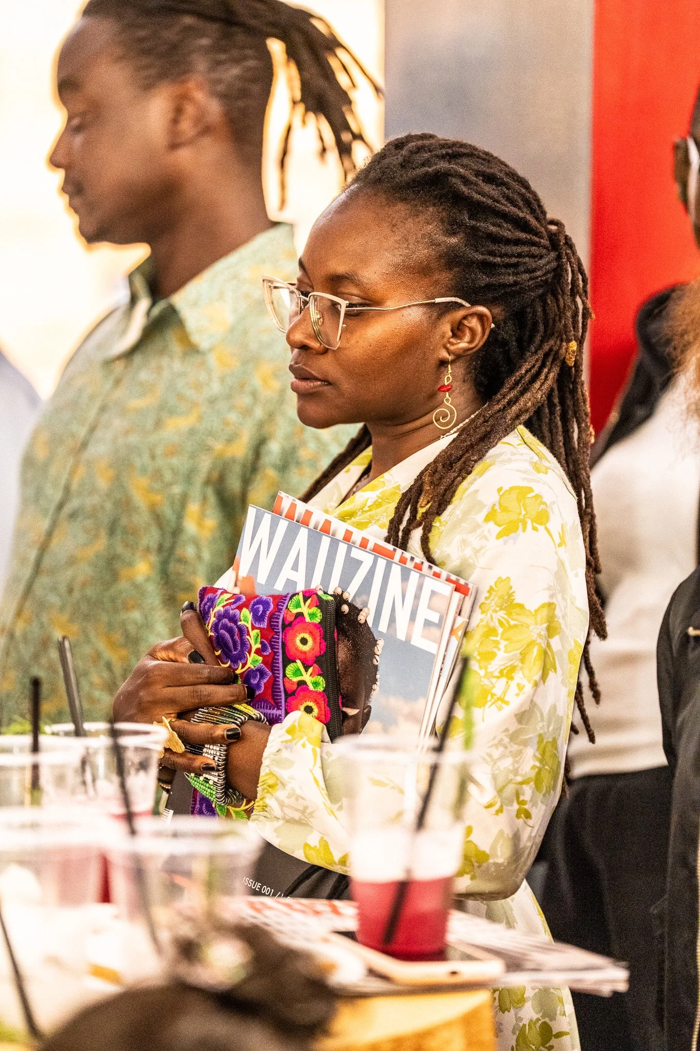 A woman with glasses and dreadlocks holding a colorful embroidered clutch and a magazine in a crowd.
