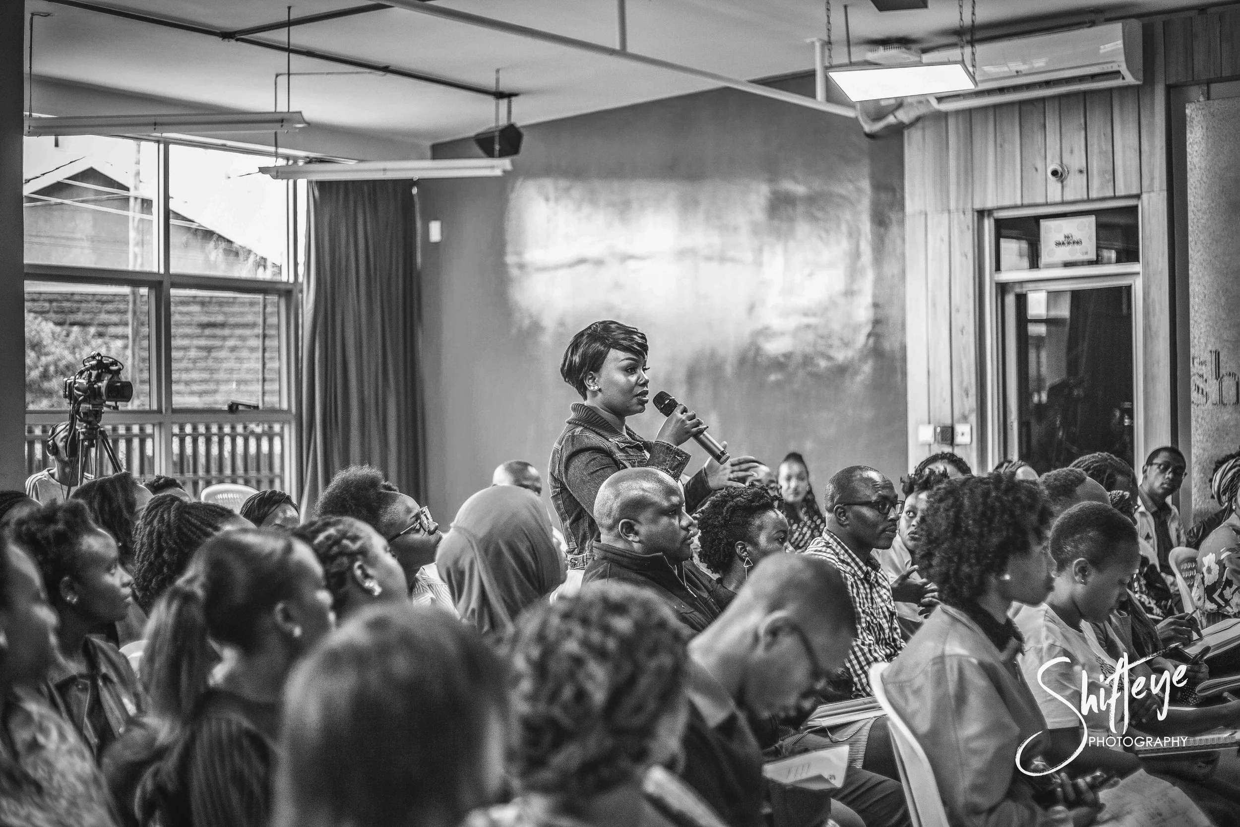 A woman stands and speaks into a microphone during a presentation or discussion in a conference room filled with seated attendees, mostly women. The room has large windows, curtains, and wooden walls.