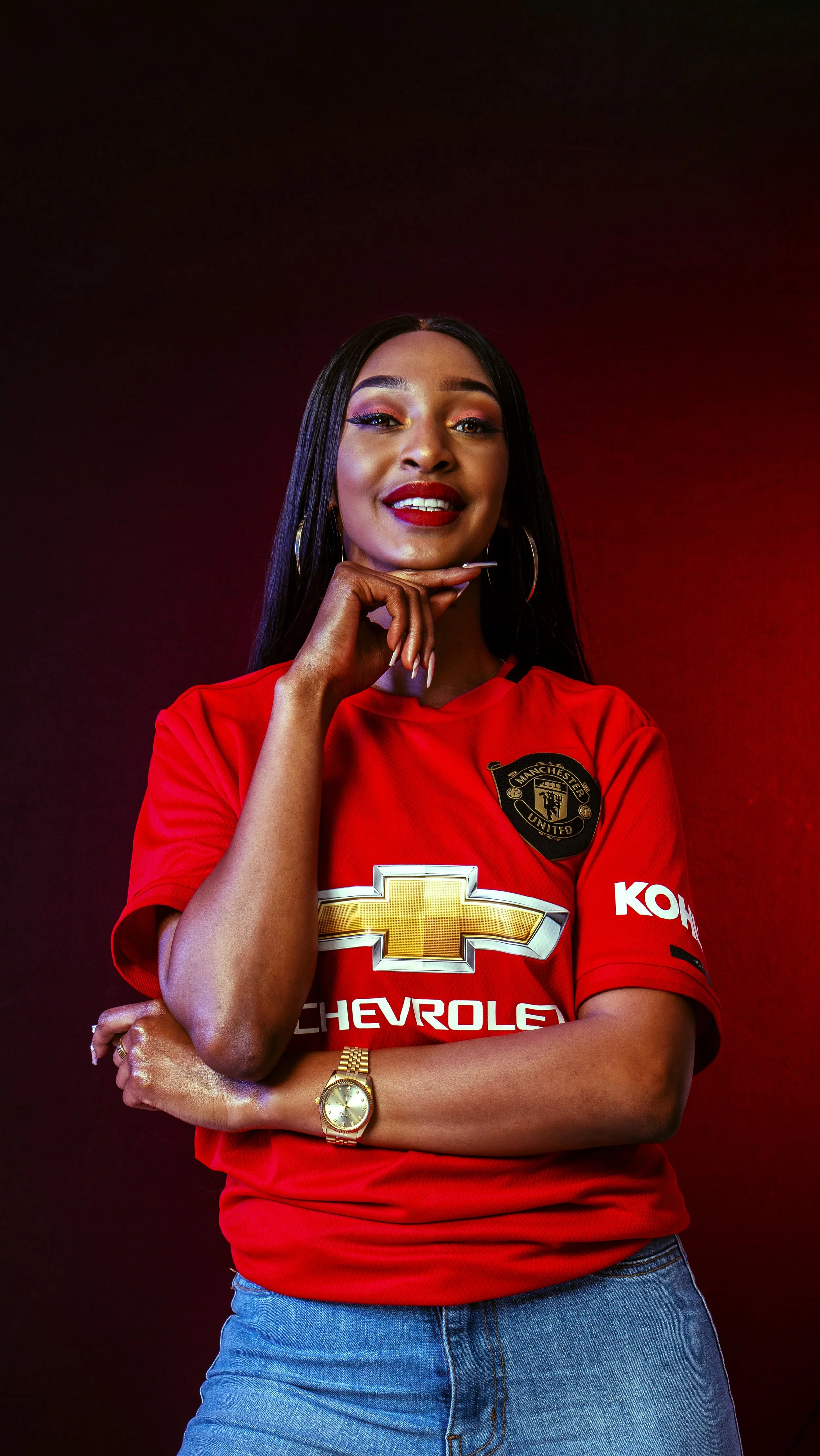 A woman smiling and posing, wearing a red Manchester United soccer jersey, gold watch, and jeans, with a dark red background.