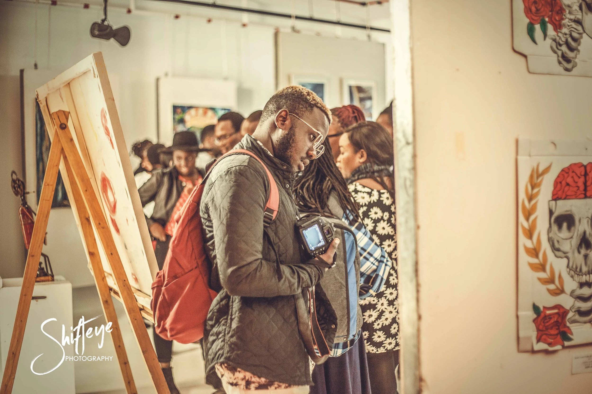 Young man with glasses and a backpack looking at his camera in an art gallery, surrounded by other people viewing artwork.