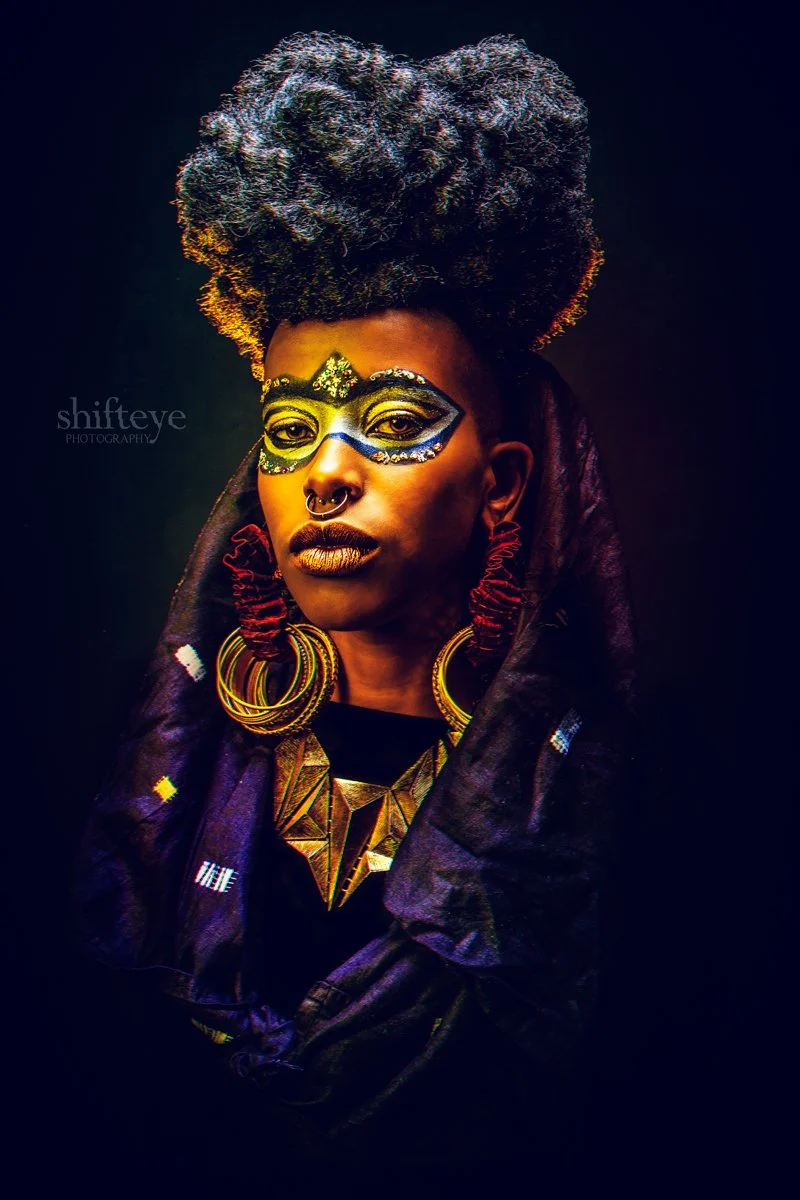 A woman with dark skin and styled gray hair wearing vibrant makeup, including a decorative face mask painted around her eyes, large gold hoop earrings, an ornate gold necklace, and a black and colorful patterned headscarf.