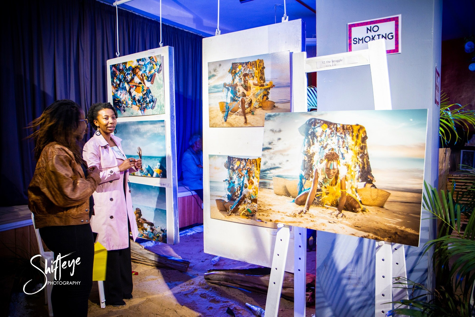 Two women viewing colorful beach-themed photographs displayed on white panels at an art exhibition or gallery, with a 'No Smoking' sign visible in the background.