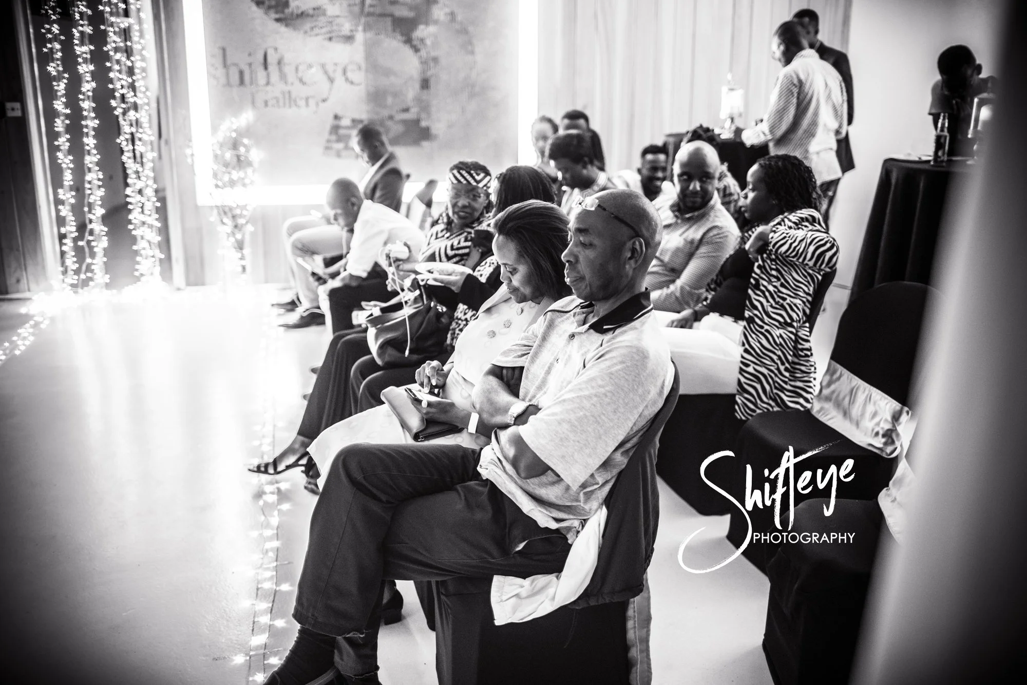 Black-and-white photo of a group of people sitting in a decorated room, attending an event or gathering, with some using their phones and others engaged in conversation.