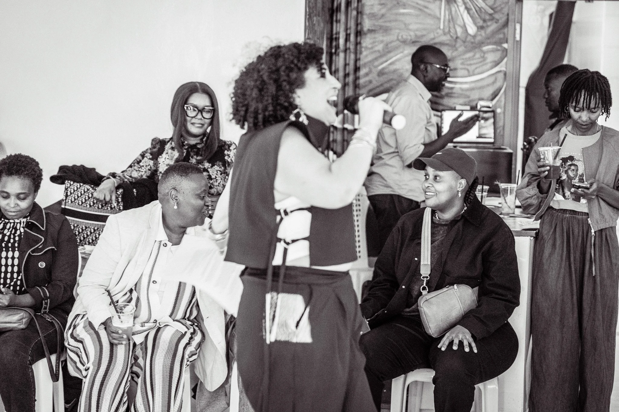 A group of women and men at a social gathering, with one woman singing into a microphone while others are listening, talking, and drinking drinks.