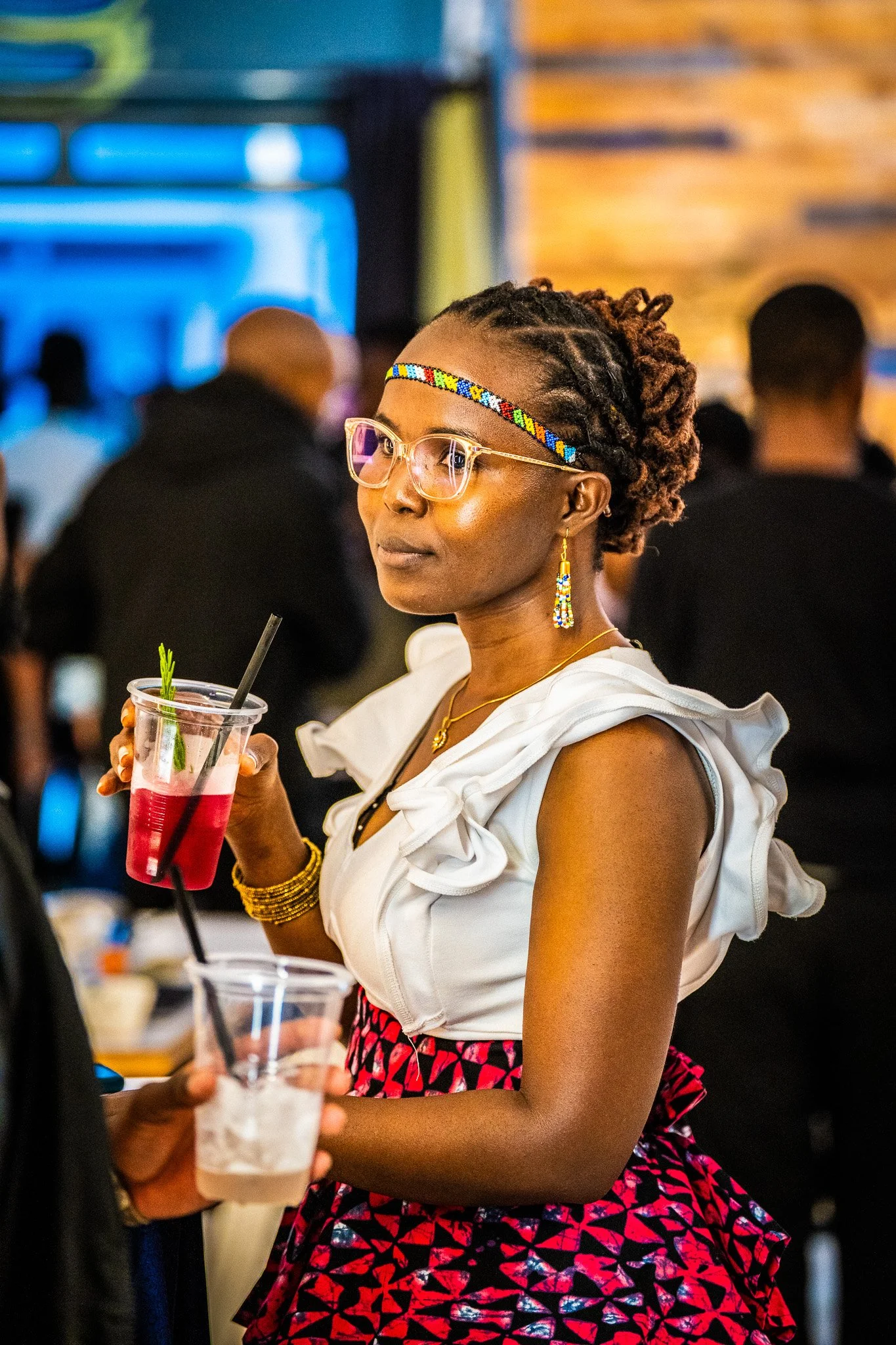 Woman with short dreadlocks wearing glasses, beaded headband, earrings, and a white ruffled top, holding a cocktail glass at a social event.