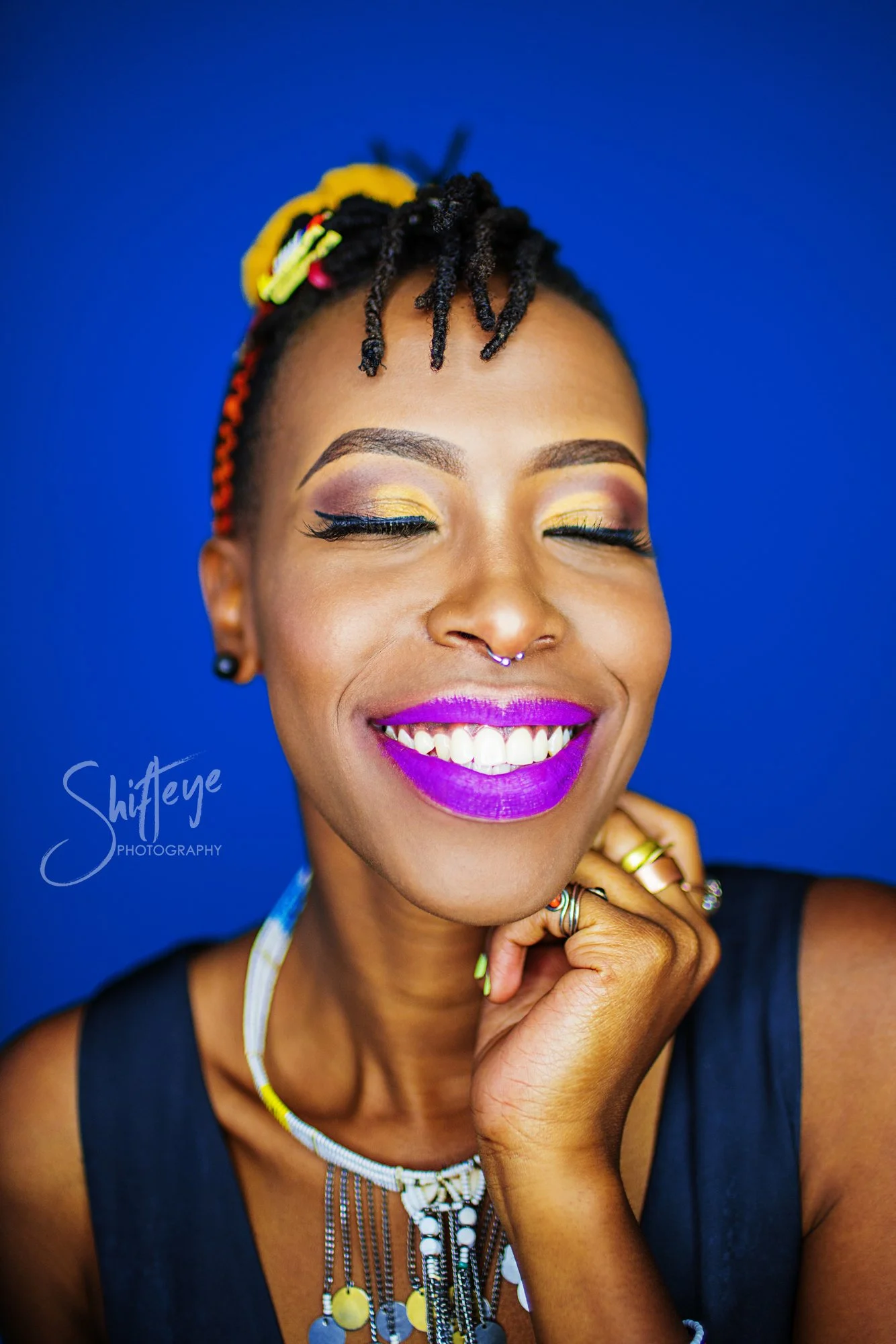 A woman with colorful makeup, bright purple lipstick, and braided hair smiling with eyes closed against a blue background, wearing jewelry and a black top.