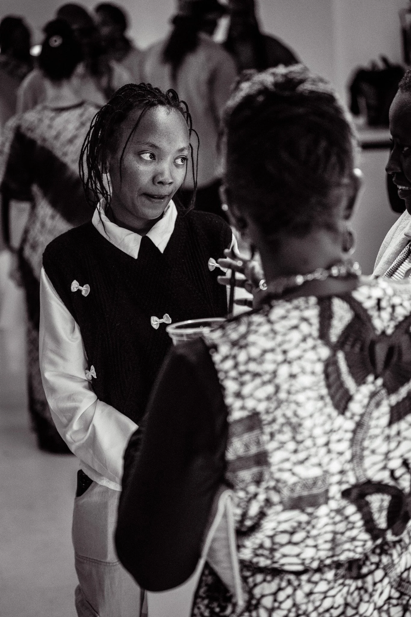 A woman with wet, braided hair speaking to a woman with short hair wearing a patterned jacket at a social gathering, with other people in the background.