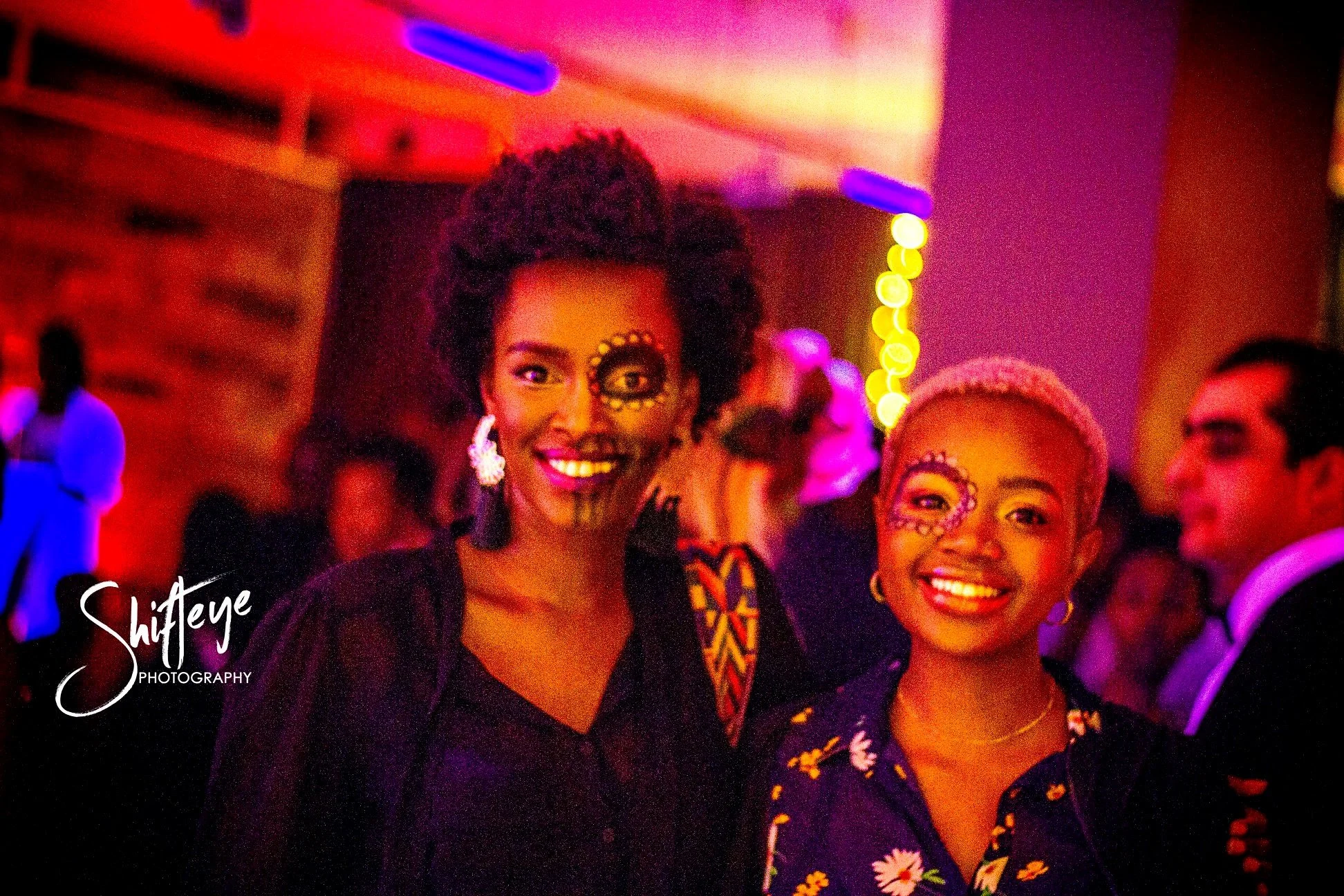 Two women with face paintings and festive attire smiling at a lively party with colorful lighting and background guests.