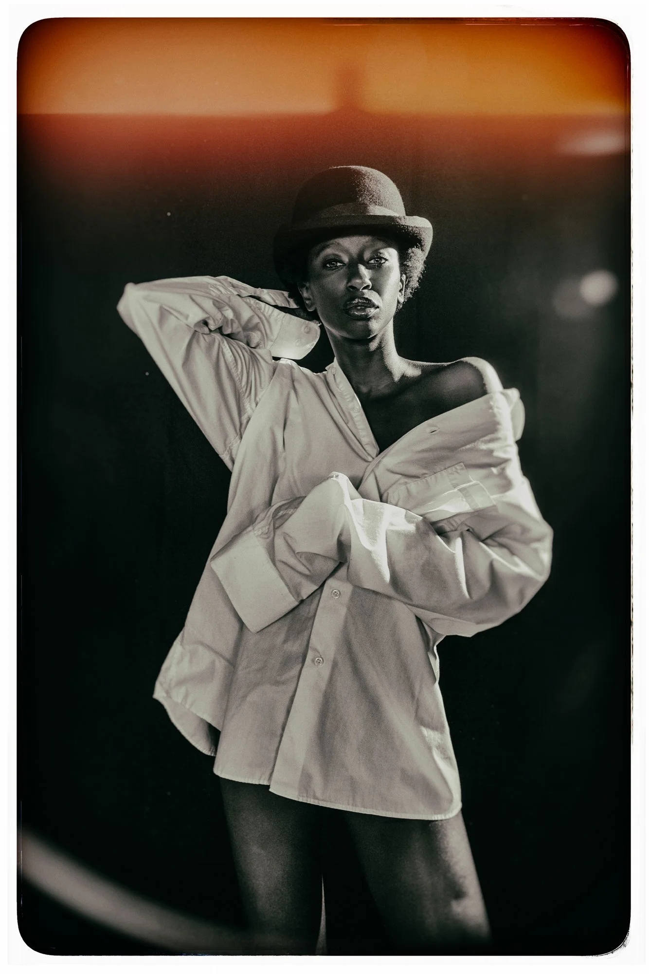 Black-and-white portrait of a woman wearing a hat and an oversized shirt, posing with one arm behind her head against a dark background with orange light at the top.