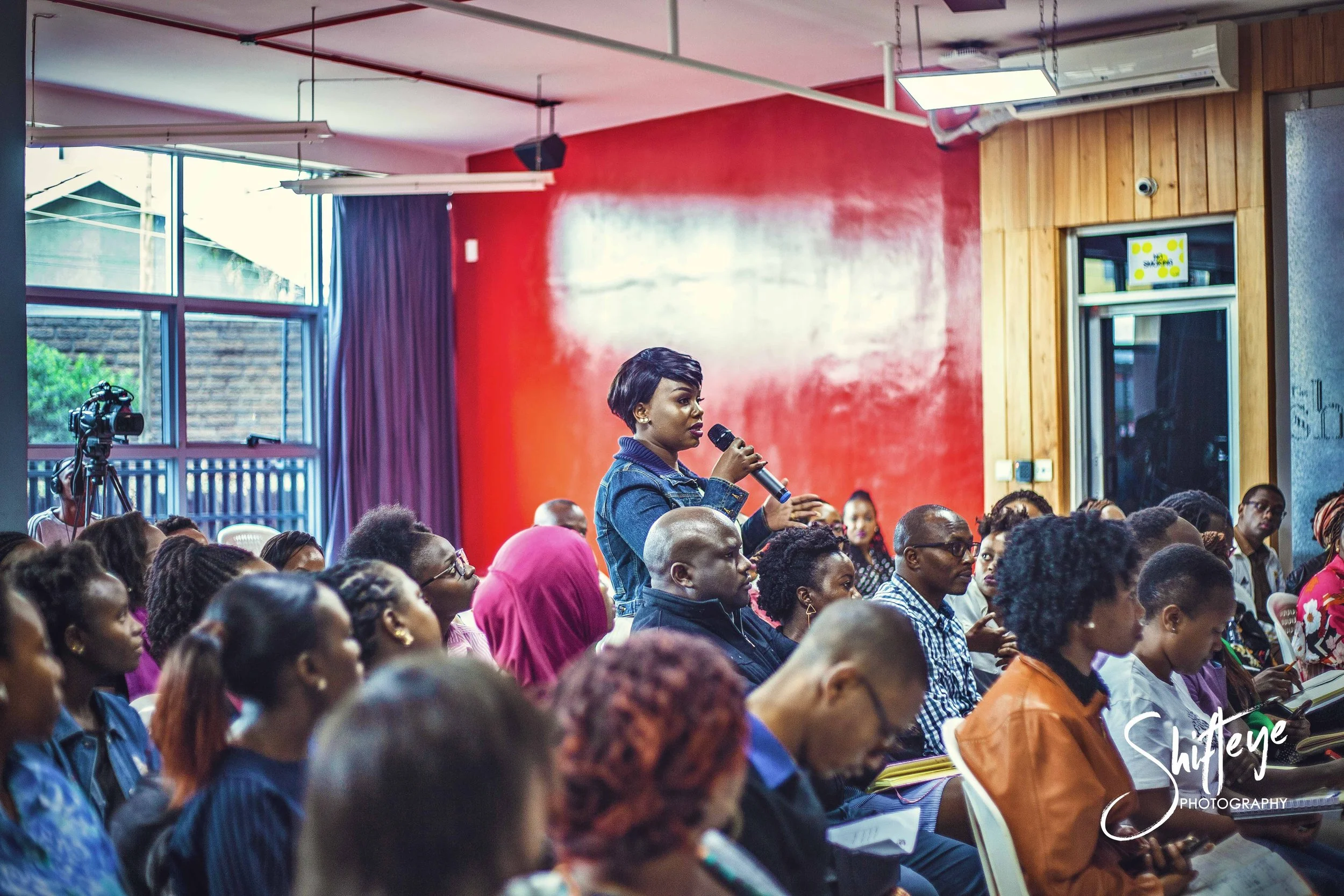 A woman standing and speaking into a handheld microphone during a conference or seminar, surrounded by seated attendees in a colorful indoor space with large windows and a mix of red, purple, and wooden walls.