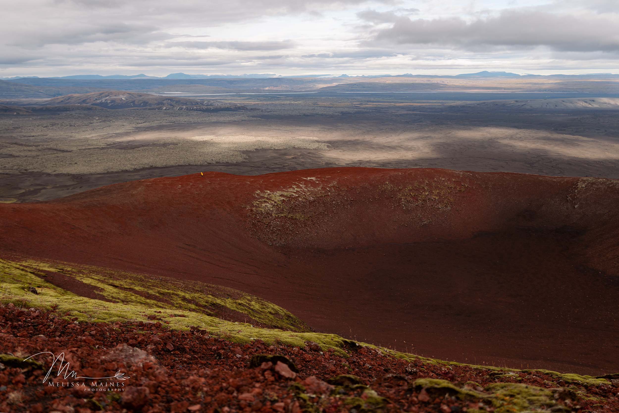 A famous Icelandic volcanic crater with red, mossy edges, overlooking a vast, desolate landscape with two people in yellow coats in the distance.