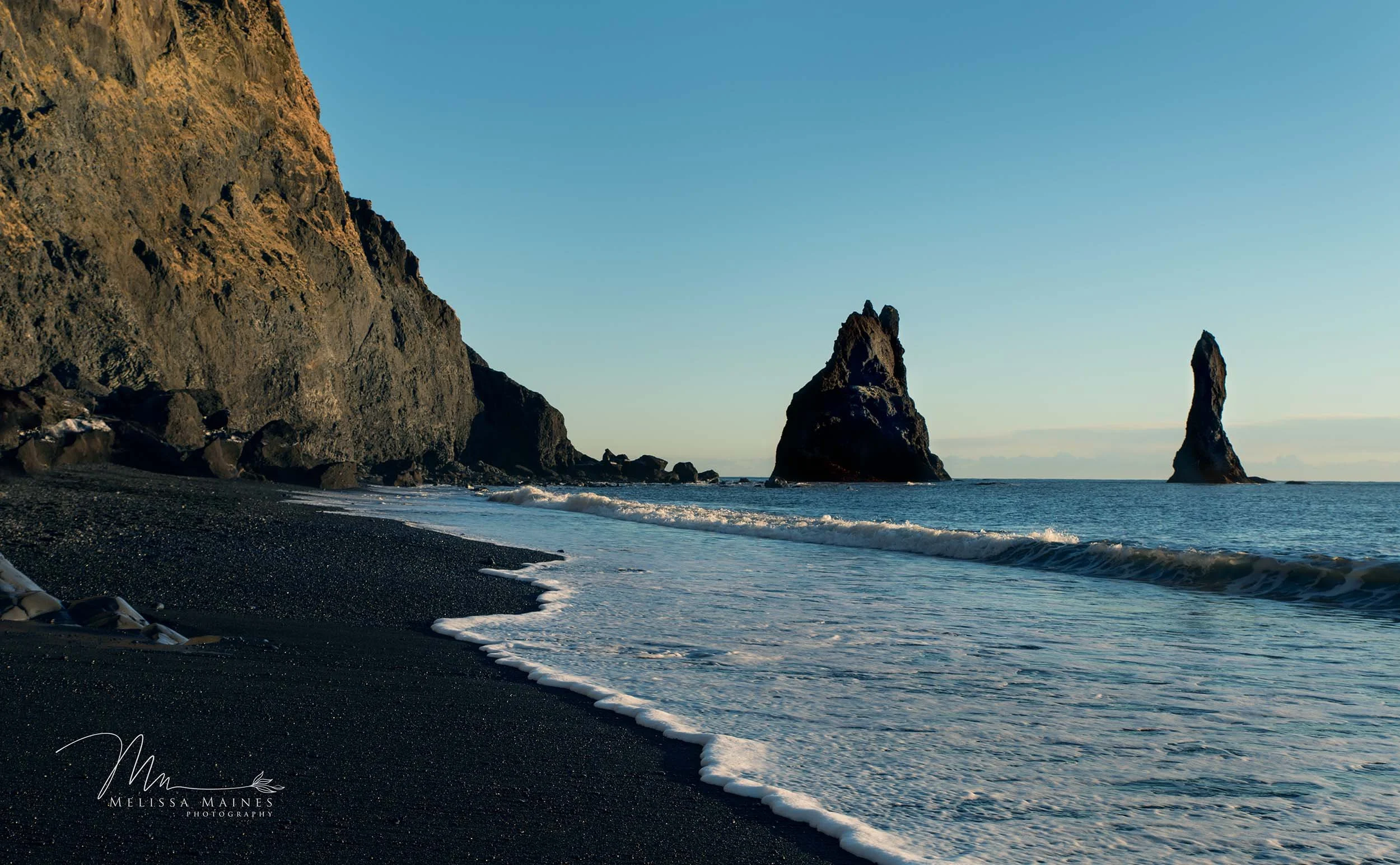Black volcanic sand beach in southern Iceland with large rock formations in the water and a cliff on the beach.
