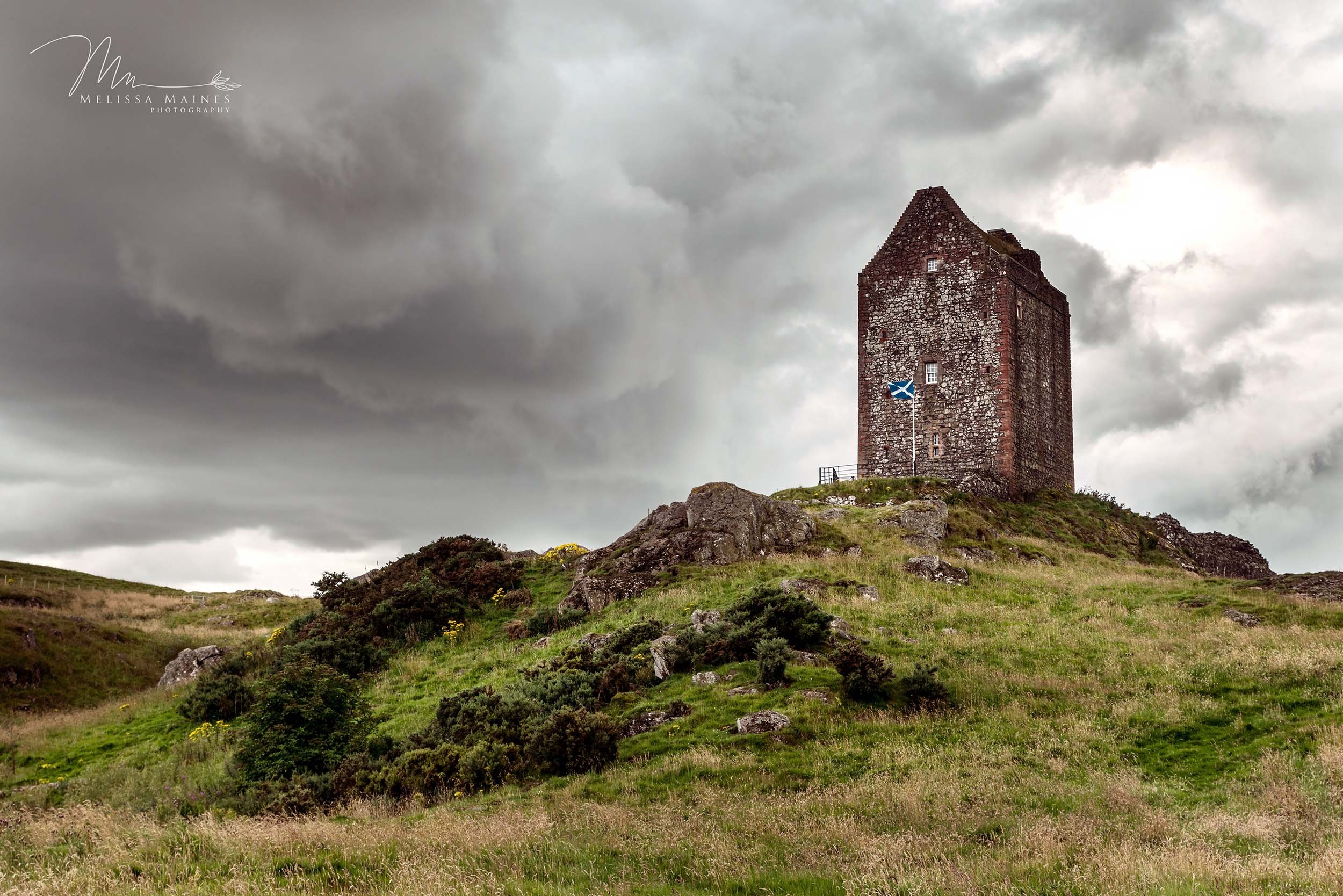 Smailholm stone tower on a grassy hill with a Scottish flag flying in front, in the border lands of Scotland.