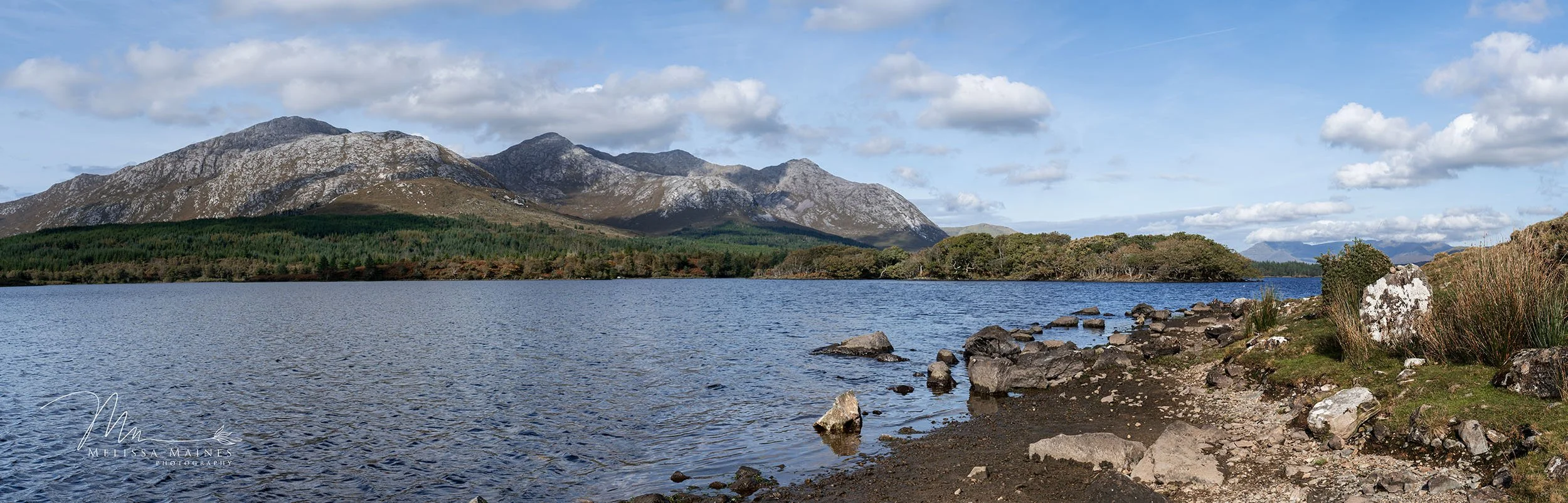 A scenic view of a lake in Ireland with rocky shoreline, surrounded by green forest and mountains.